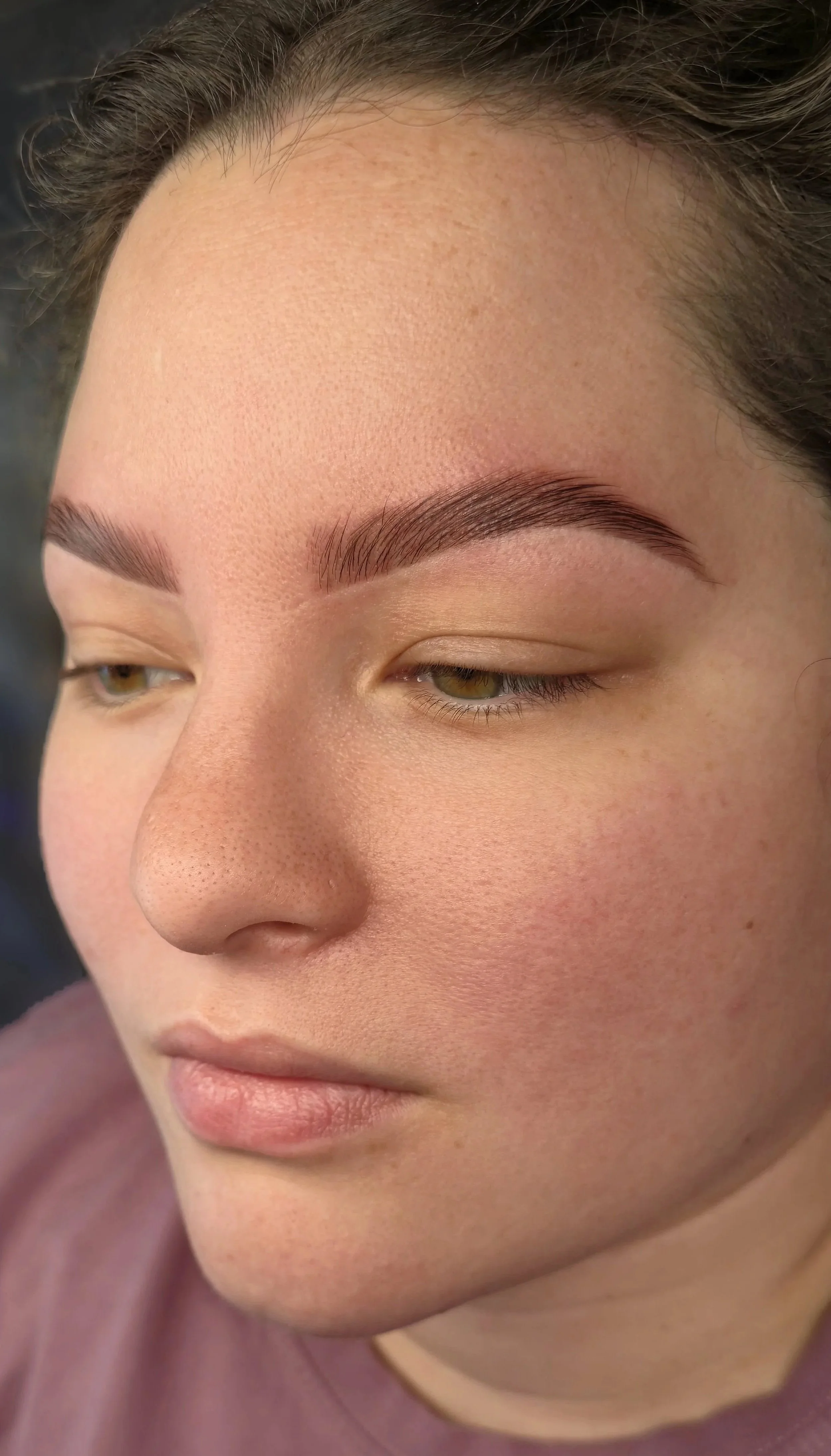 A close-up of a woman's face showing fresh eyebrow makeup, natural skin with freckles, and light lipstick, with her eyes partially closed.