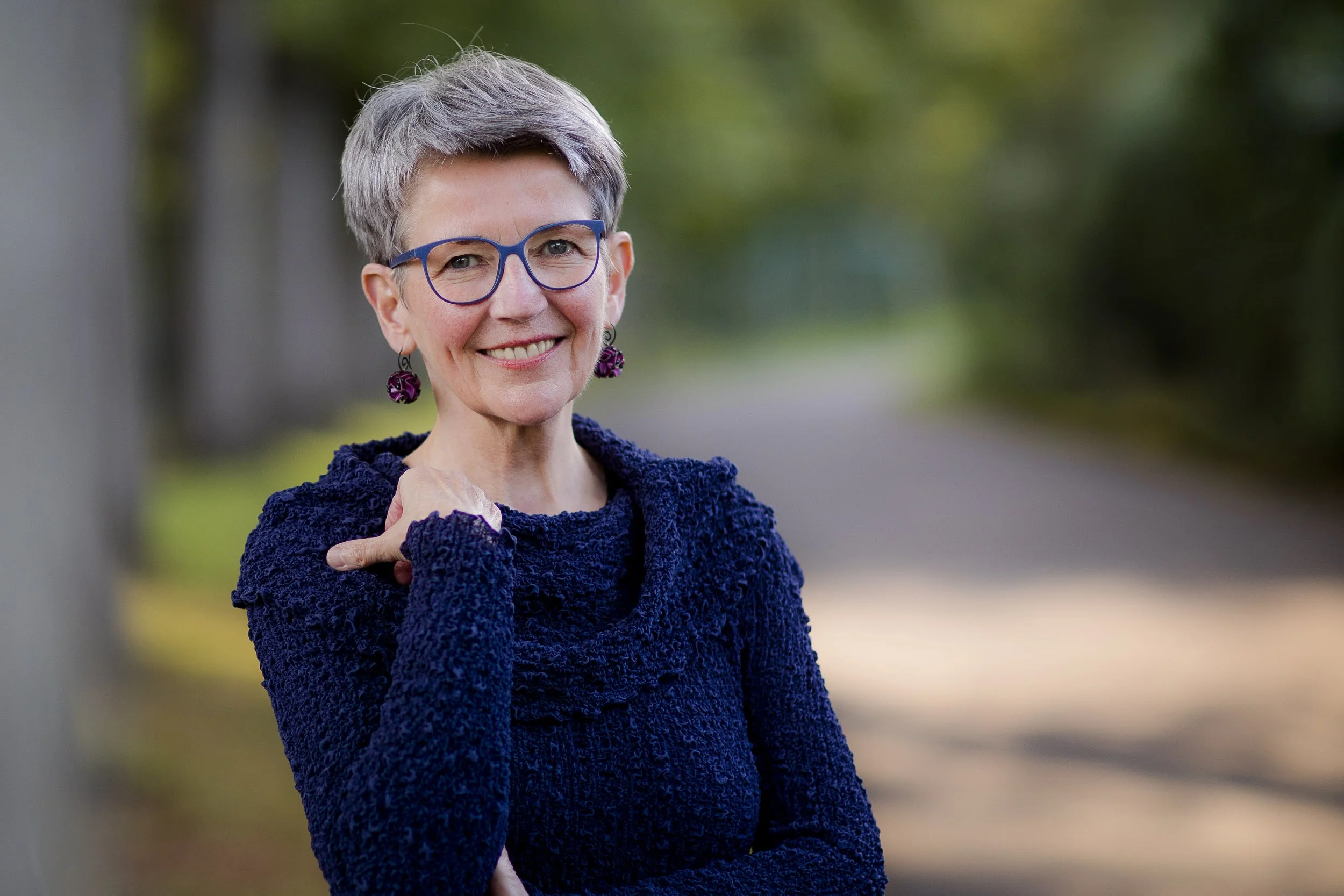 Hilde Janich mit kurzen grauen Haaren, blauen Brillen und lila Ohrringen, trägt einen dunkelblauen Pullover, steht draußen auf einem Weg mit Bäumen im Hintergrund.
