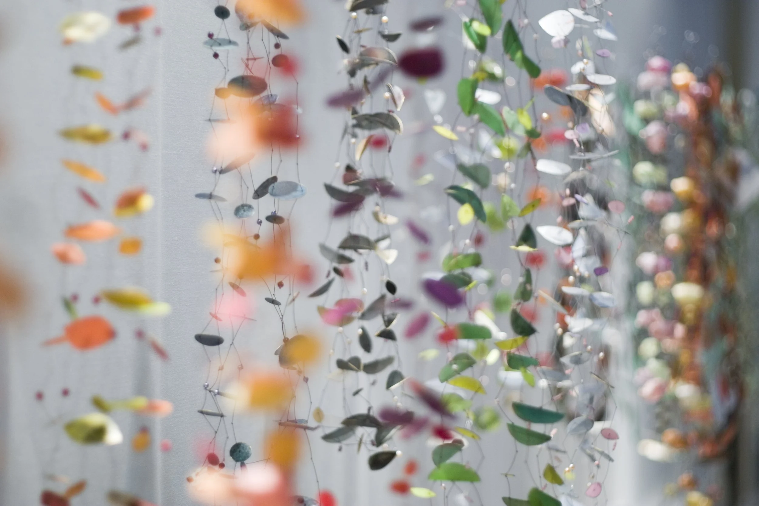 Bunte Ketten mit bunten, flachen, ovalen und runden Elementen, die an dünnen Drähten hängen, vor einem hellen Hintergrund.