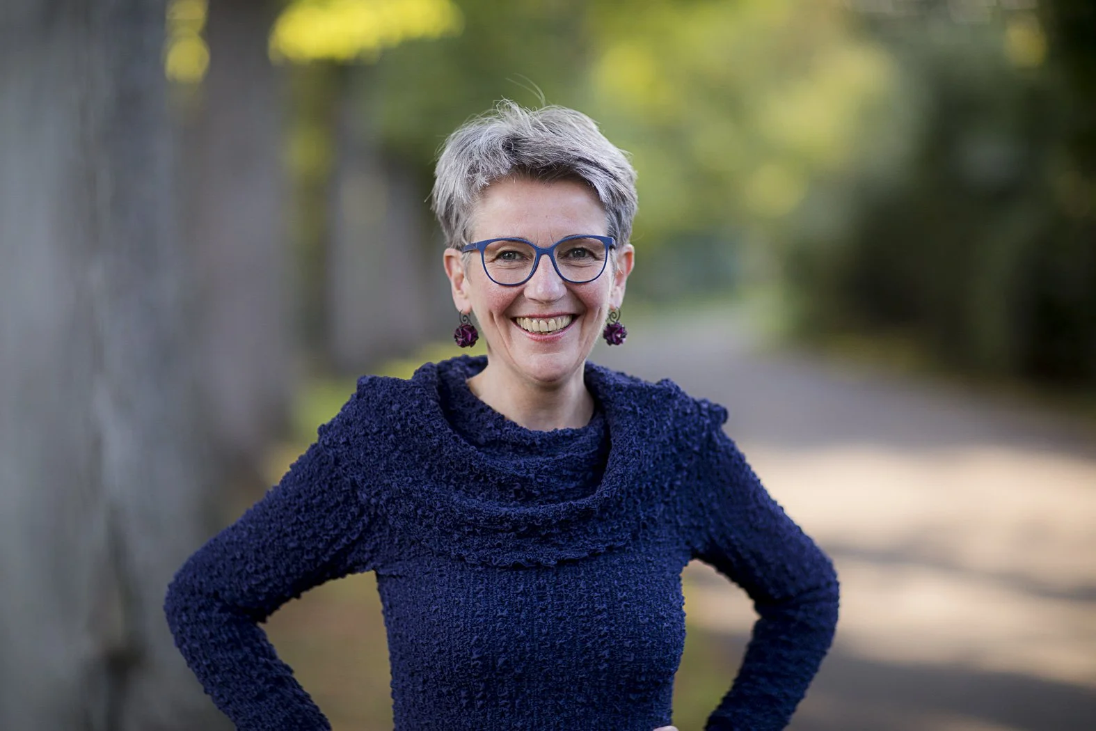 Hilde Janich mit kurzen grauen Haaren, blauen Brillen und dunklem blauen Pullover steht im Freien auf einem Weg mit grünem Laub im Hintergrund.