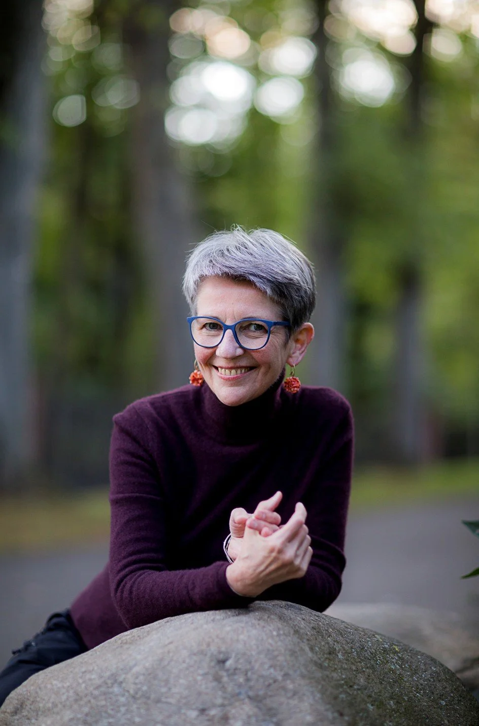Hilde Janich mit kurzen grauen Haaren, blauen Brille und roten Ohrringen lehnt auf einem großen Stein in einem Wald, lächelt und schaut in die Kamera.