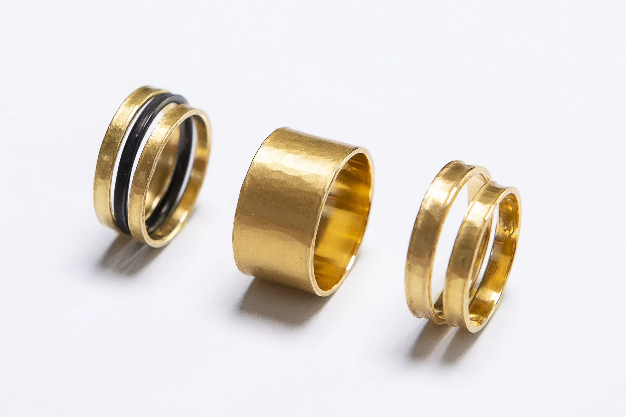 Drei goldene Ringe auf weißem Hintergrund, einer mit schwarzen Akzenten, der mittlere ist breit, der rechte sind schmal, alles aus Metall gefertigt.