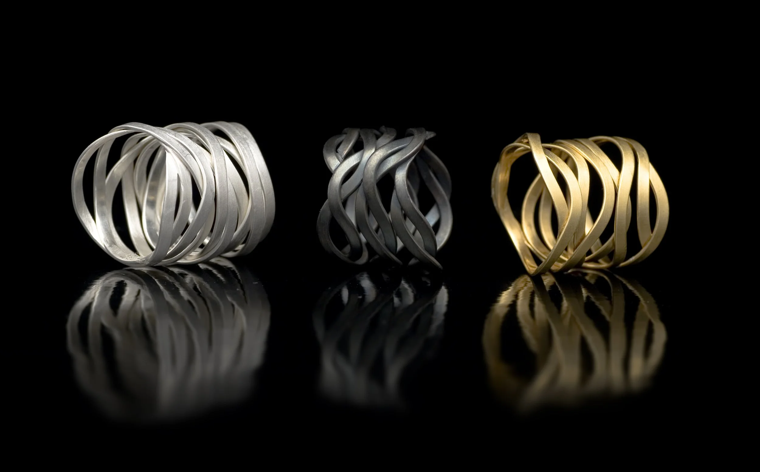 Drei verschiedenfarbige, kunstvoll gewundene Metallringe auf schwarzem Hintergrund, mit reflektierenden Oberflächen.