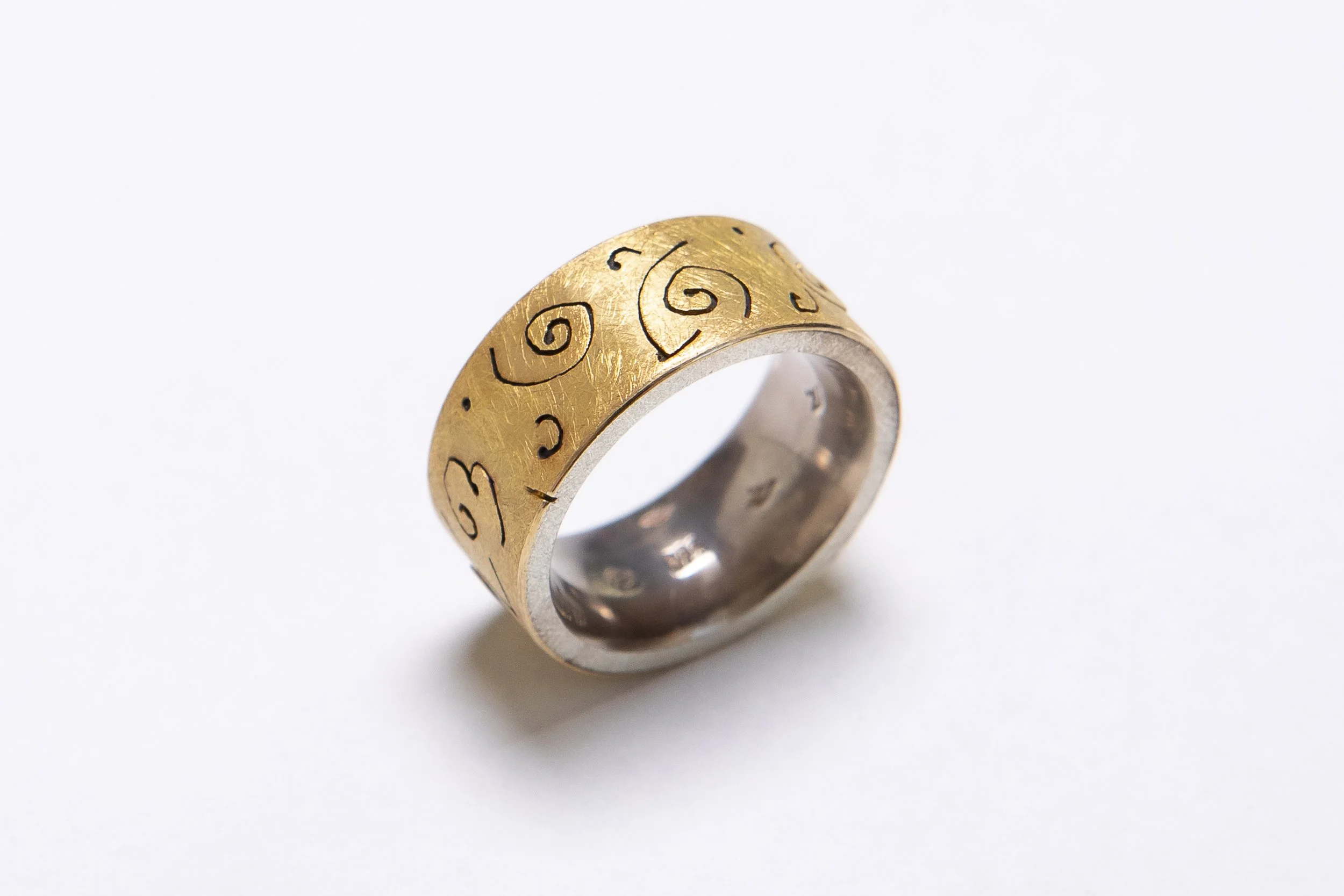 Ring mit goldener Außenseite und schwarzen Mustern, silberner Innenseite, auf weißem Hintergrund.