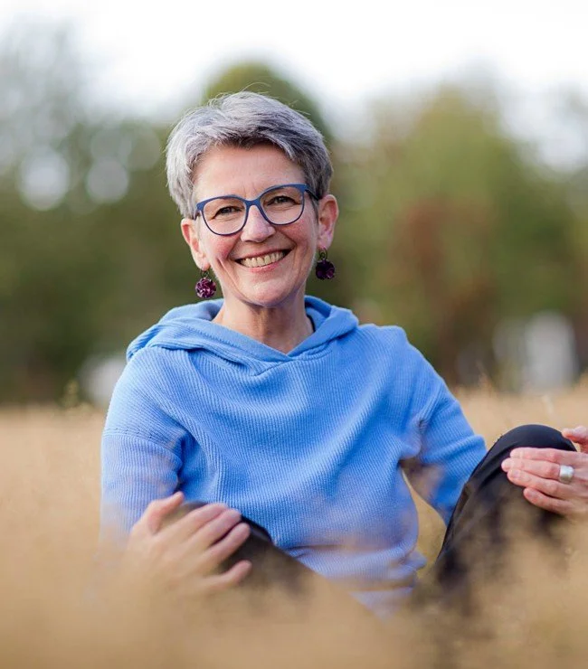 Eine lächelnde ältere Frau mit kurzen grauen Haaren, Brille und Ohrringen sitzt im Freien in einer Wiese, trägt eine hellblaues Sweatshirt.