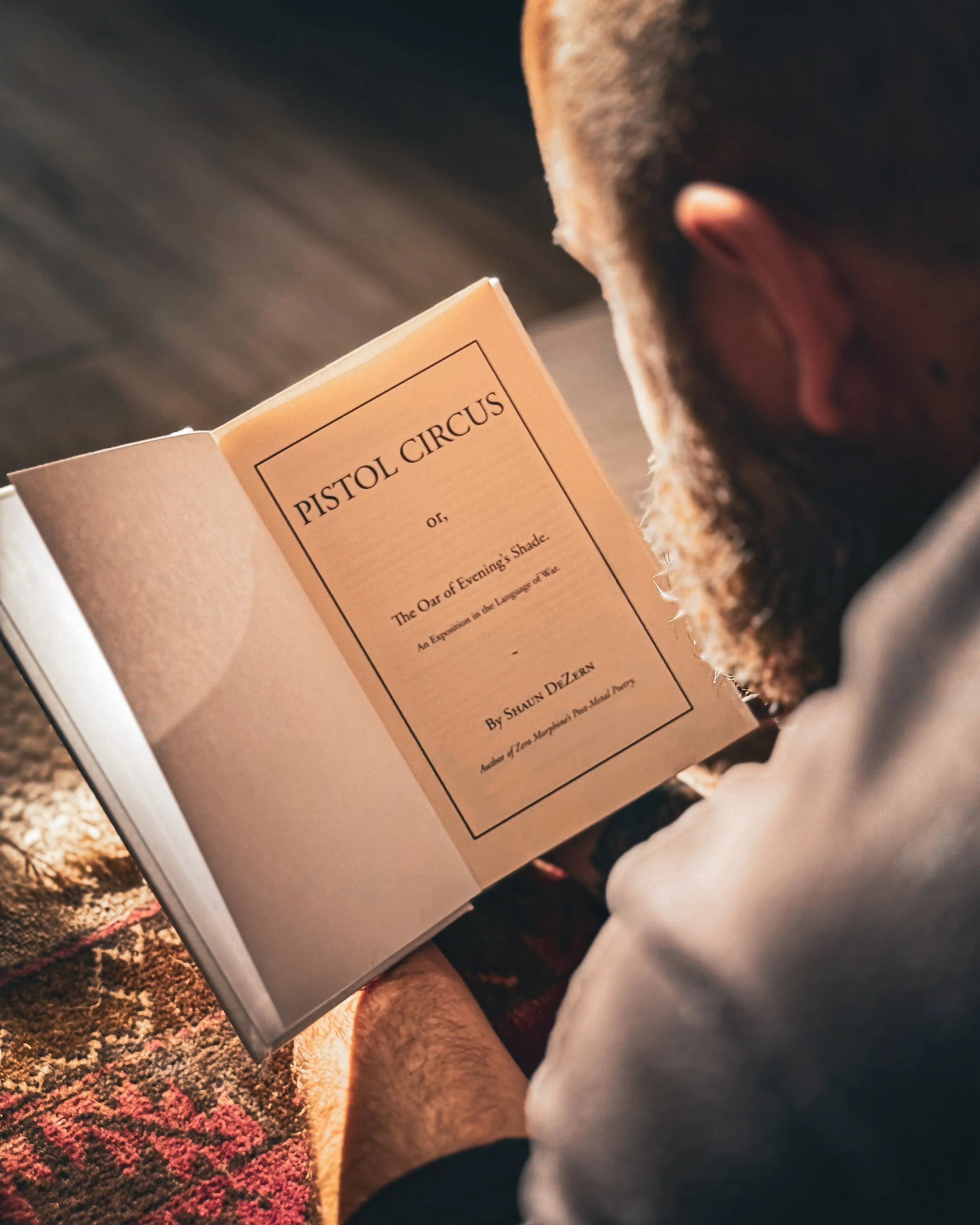 Person with a beard reading a booklet titled 'Pistol Circus' while sitting on a patterned rug.