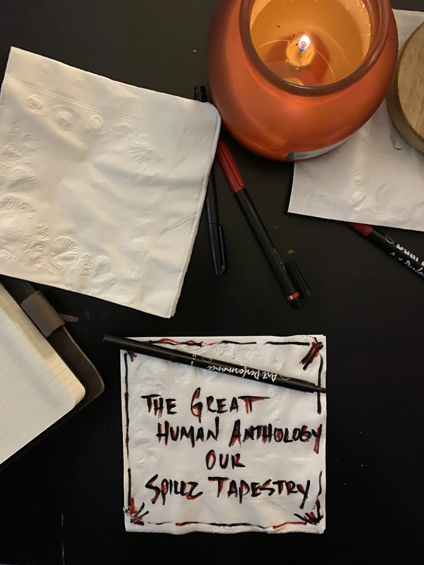 A protest sign with red and black writing that says, "The Great Human Anthropology Dump Our Squizz Tapestry." There is a candle in an orange holder, a black marker, and a notebook with lined paper on a dark surface.