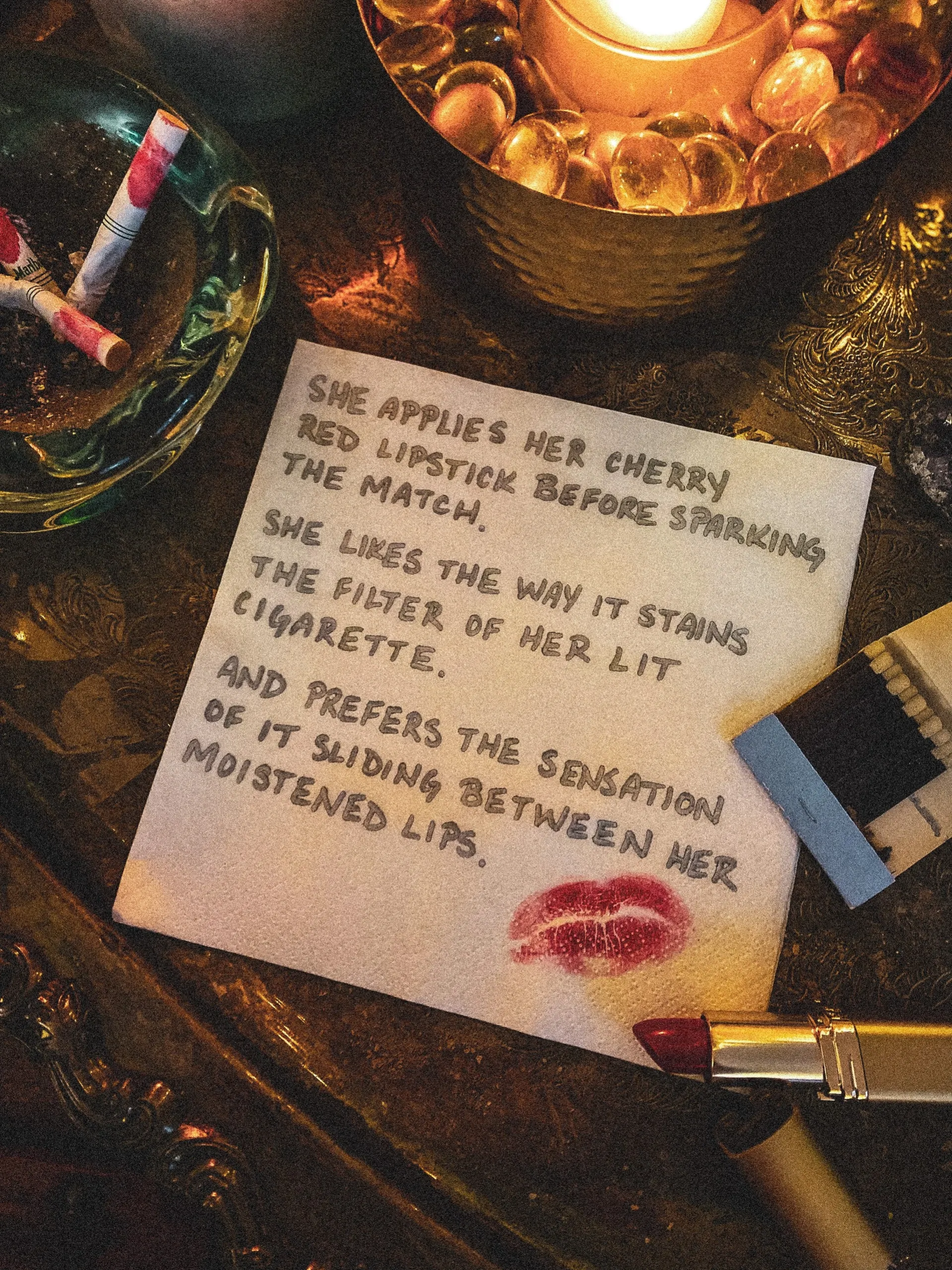 A handwritten note with a red lipstick kiss mark on it, placed on a dark wooden table. The note describes a woman applying her cherry red lipstick before sparking the match and liking the way it stains her lips, with a lipstick and a pen nearby. There is a lit candle in a decorative holder and a small bowl with glass marbles on the table.