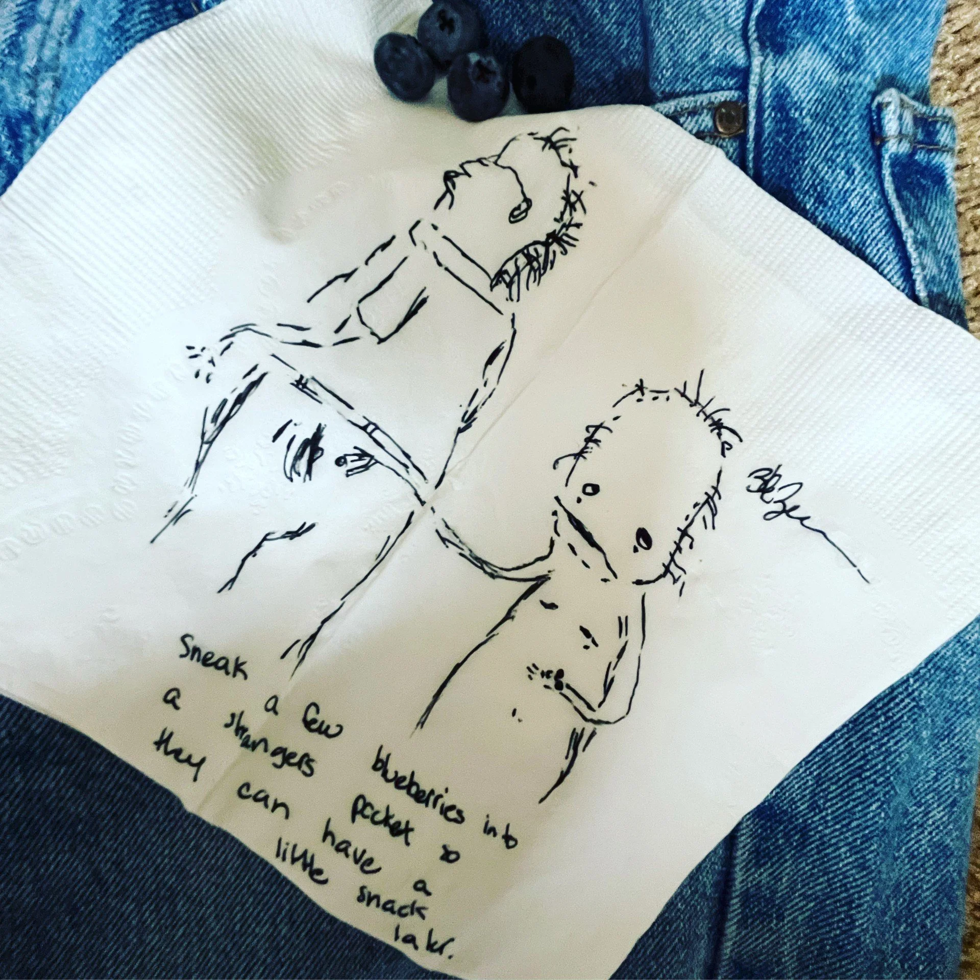Close-up of a handwritten letter with a sketch of a girl and a dachshund, a small cluster of blueberries, and part of a denim jacket and a piece of textured fabric in the background.