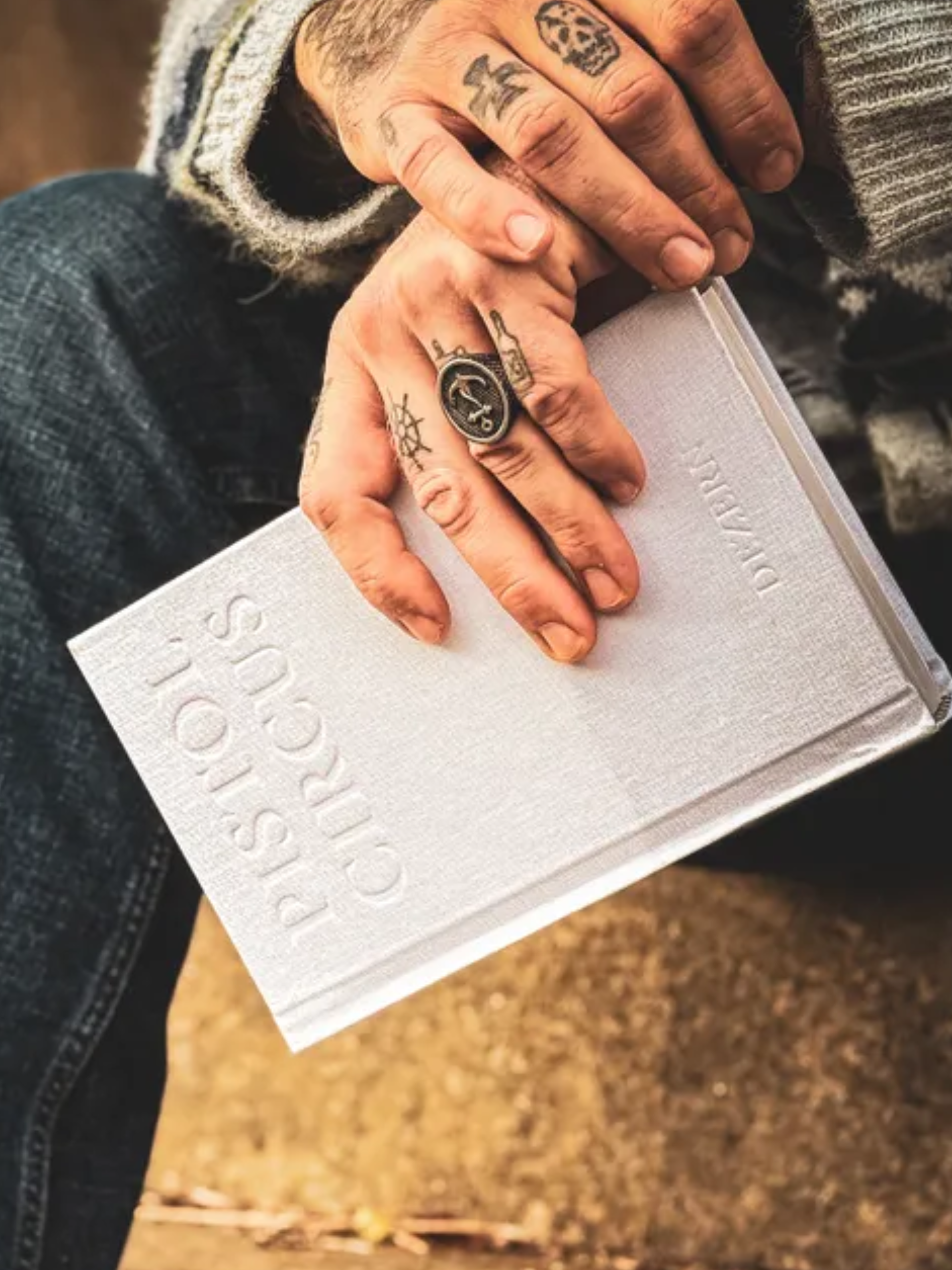 Close-up of a person’s hand with multiple tattoos, holding a white hardcover book. The person is wearing a gray and white striped sleeve, and the background features a brown, speckled surface.