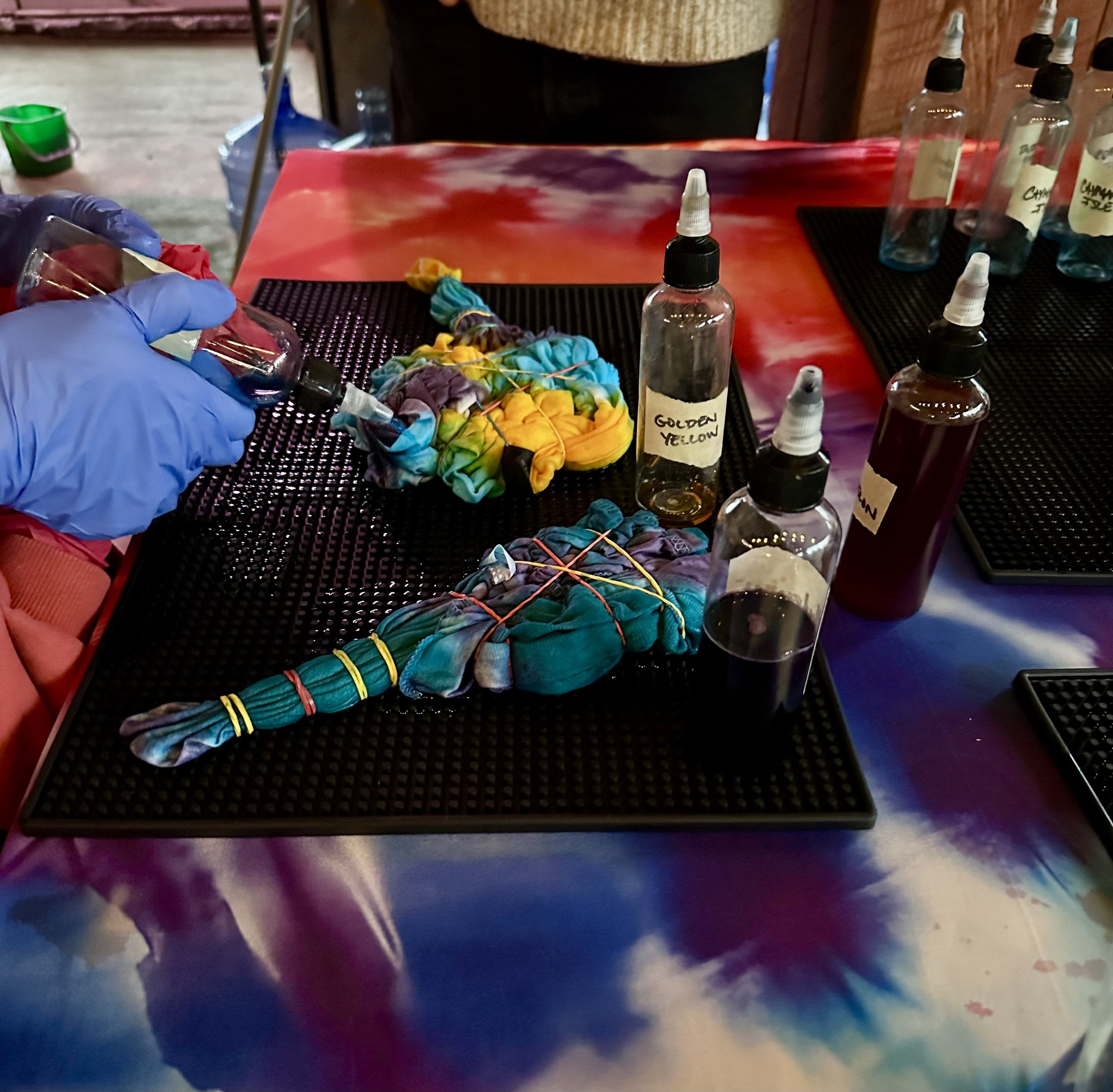 A person wearing blue gloves using a pipette to transfer liquid into a small bottle labeled 'GOLDEN YELLOW'. The table has several small bottles with droppers and some colorful fabric or yarn, and the background shows a wooden wall.