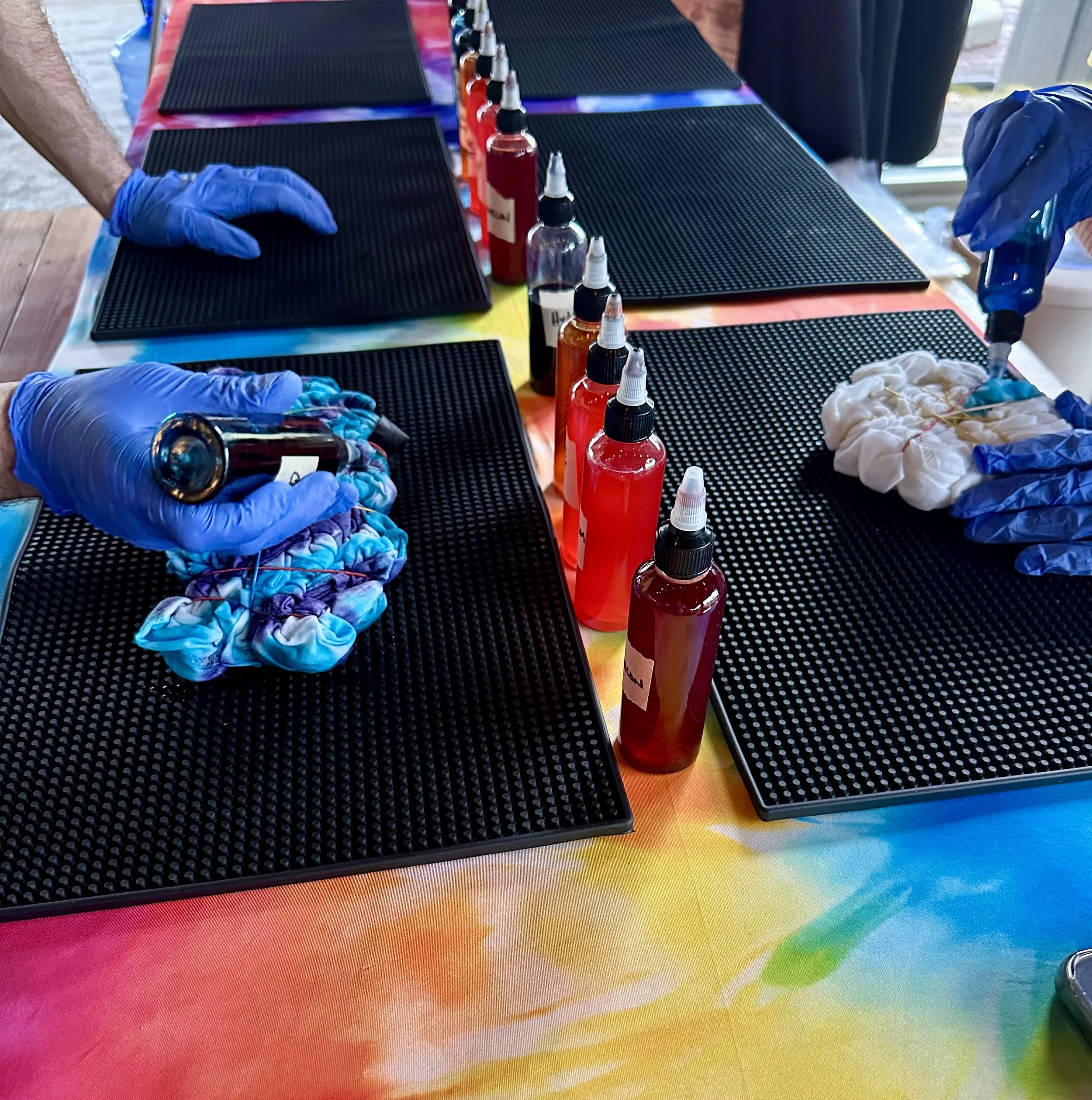 A table with black silicone mats, bottles of tattoo ink, a tattoo machine, and gloves, set on a colorful rainbow tablecloth.