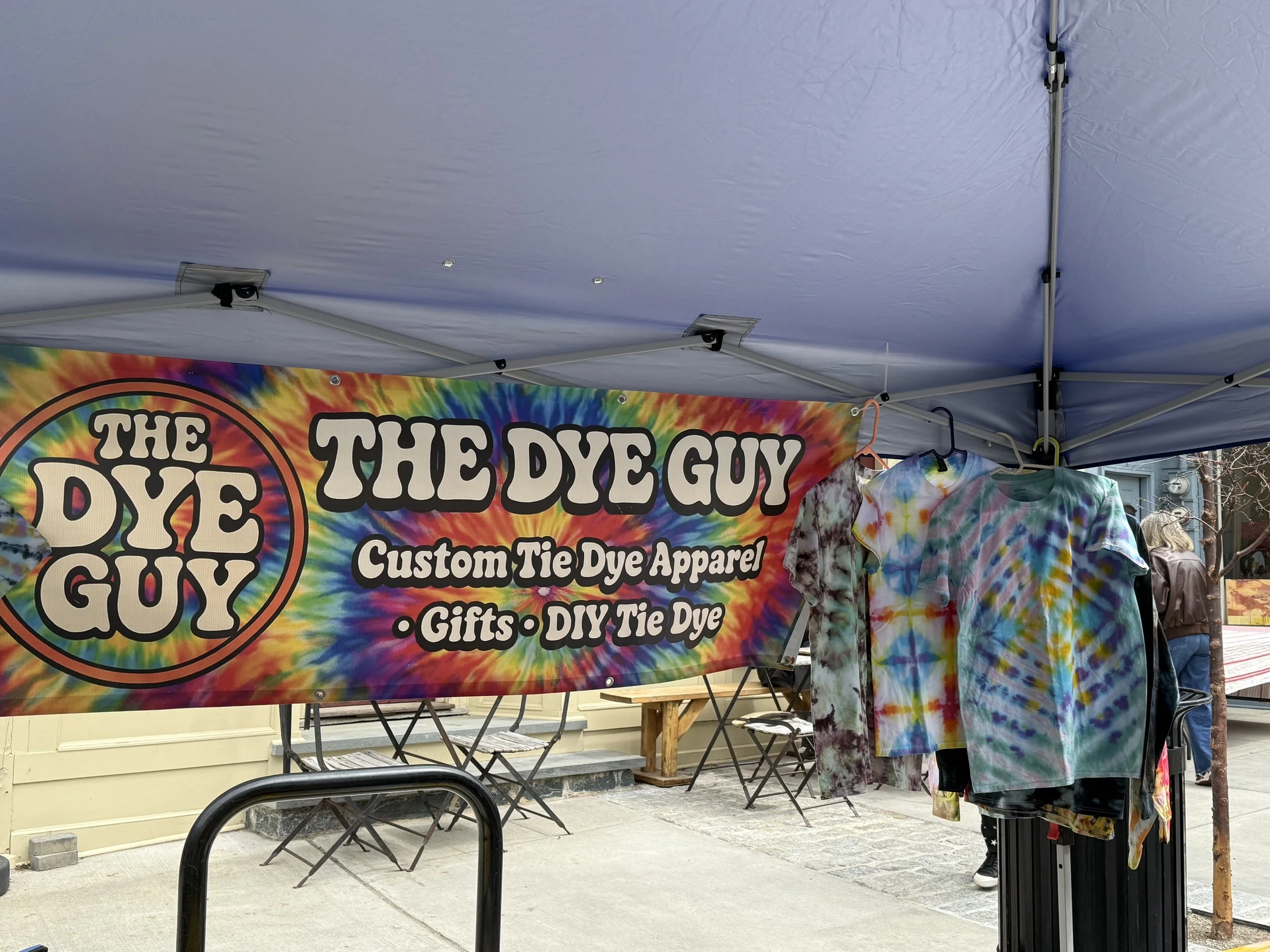 A colorful tie-dye banner advertising The Dye Guy for custom tie-dye apparel, gifts, and DIY tie-dye services, with tie-dye shirts hanging in front of the banner at an outdoor event.