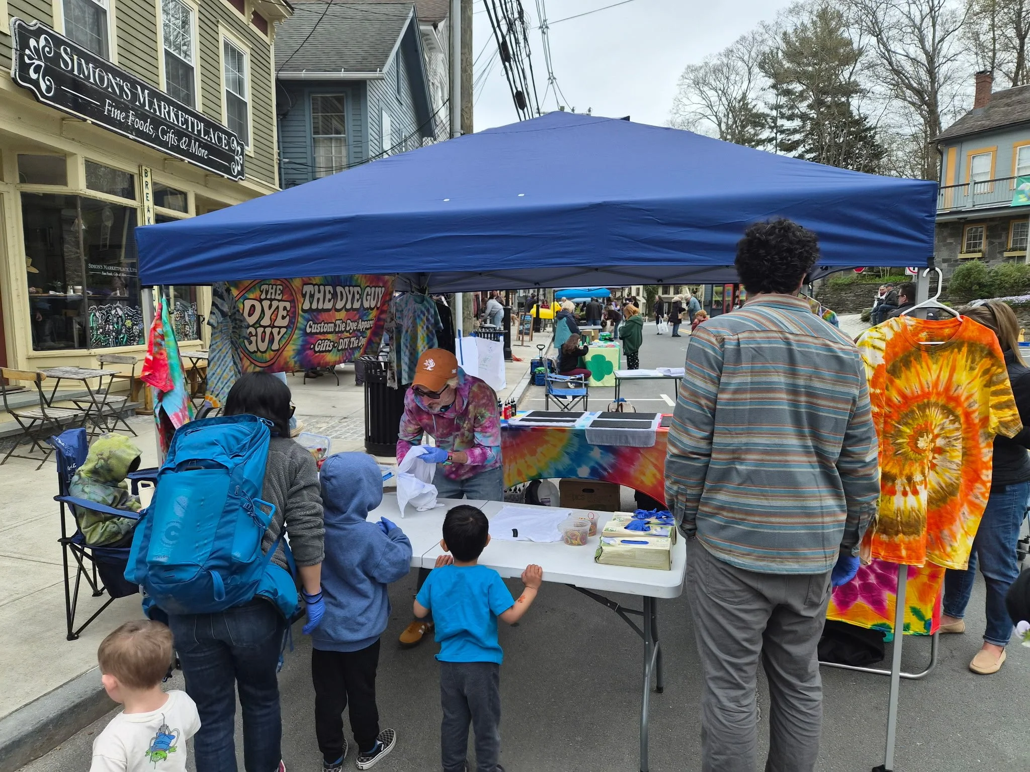 People gathered at an outdoor market stall with a blue canopy, selling tie-dye shirts and crafts.