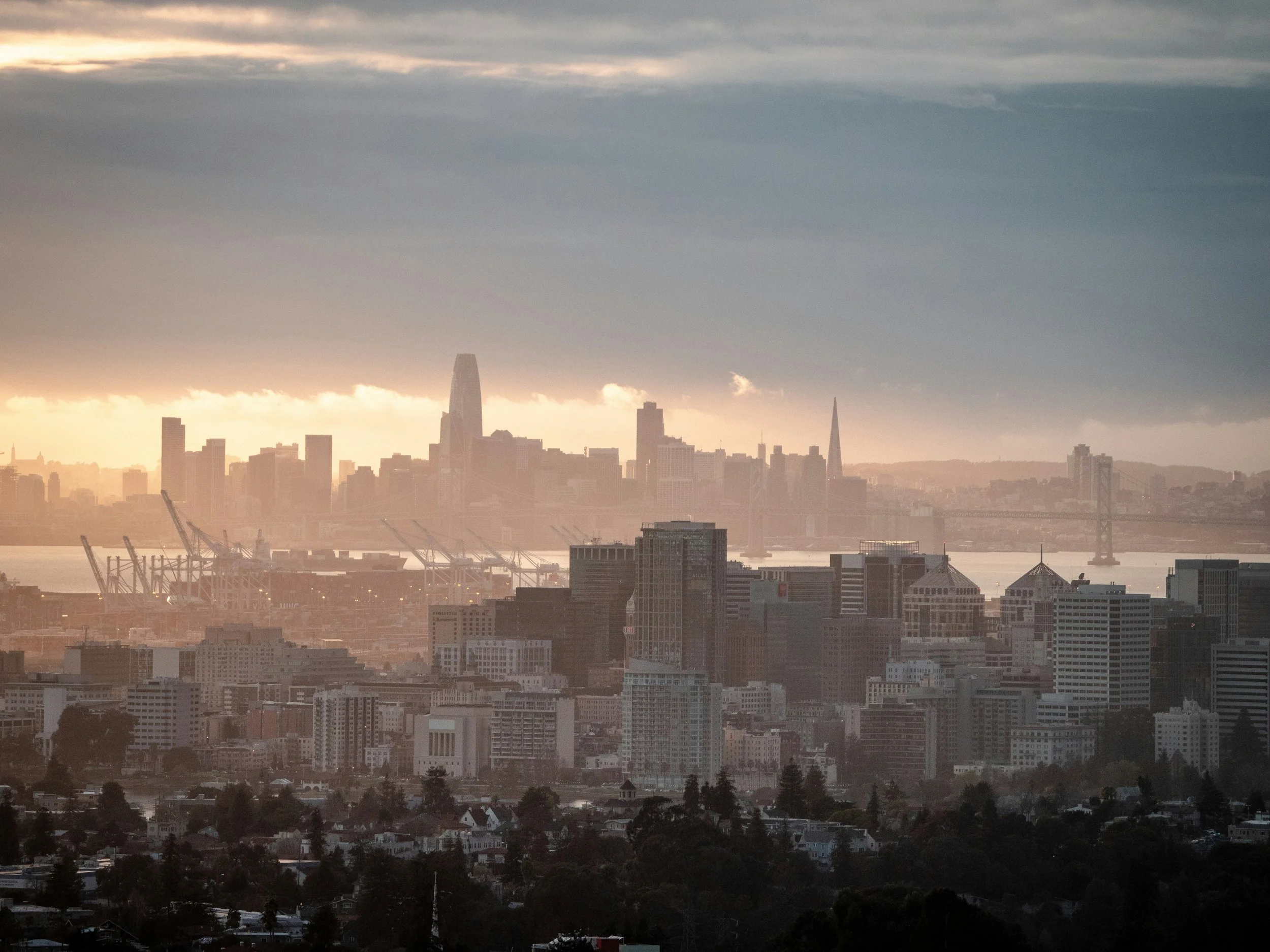 City skyline with tall buildings and bridges, hazy sunset over water in Oakland and San Francisco.