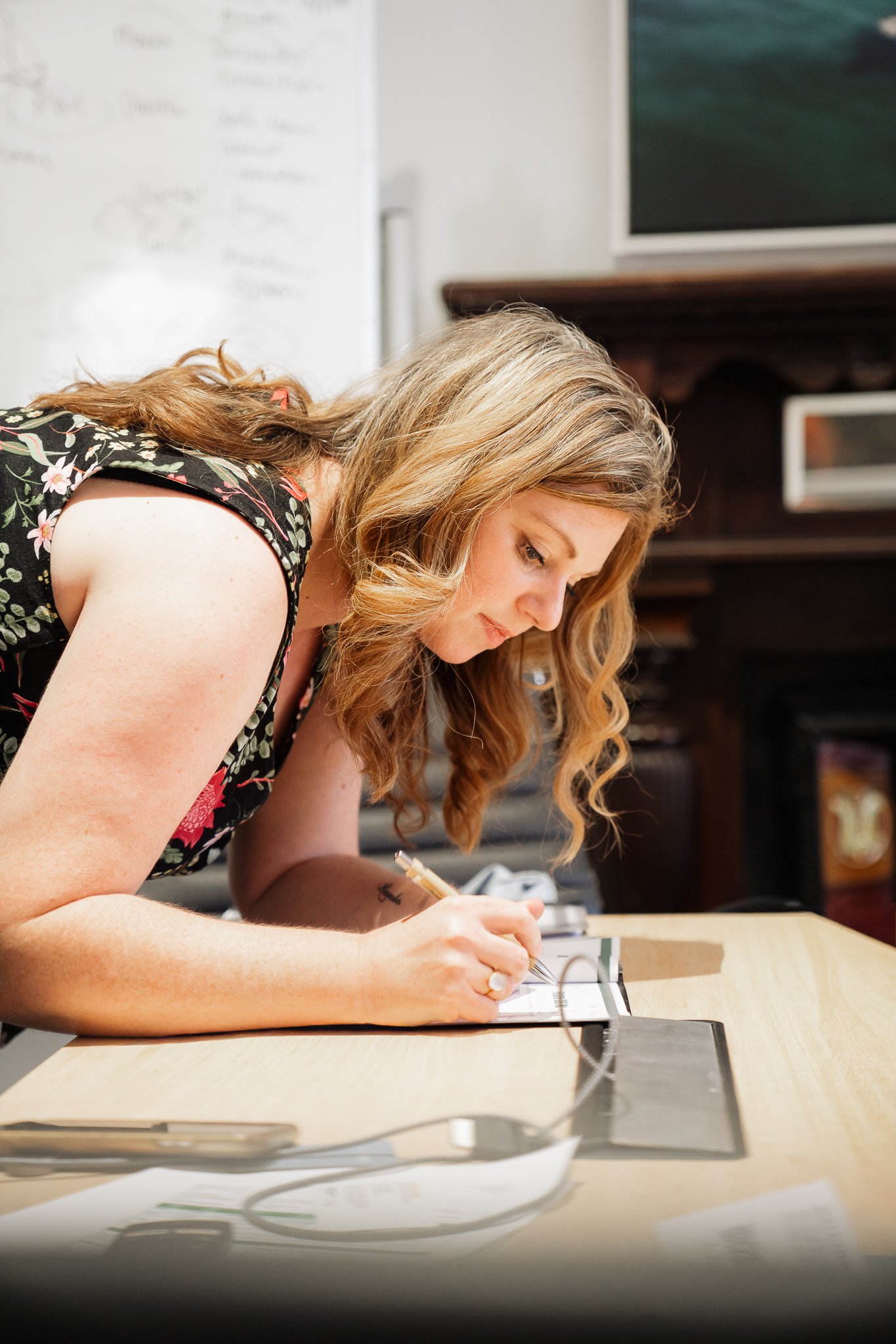 A woman with wavy, shoulder-length blonde hair leaning over a wooden table, writing on a notepad with a pen. She is wearing a sleeveless black dress with floral patterns.