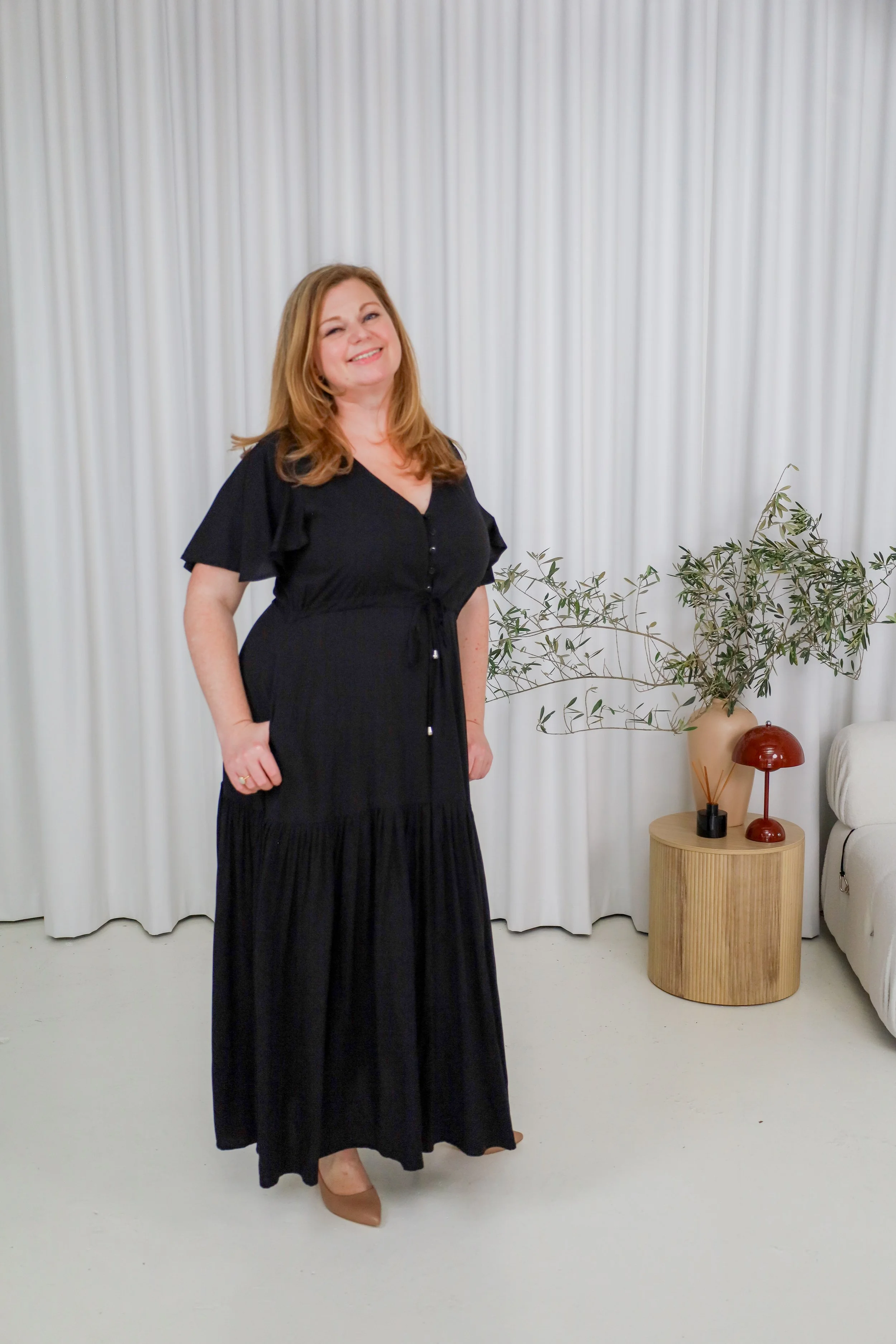 A woman with red hair, wearing a black dress and nude heels, smiling indoors with white curtains, a wooden side table, a potted plant, and decorative items.