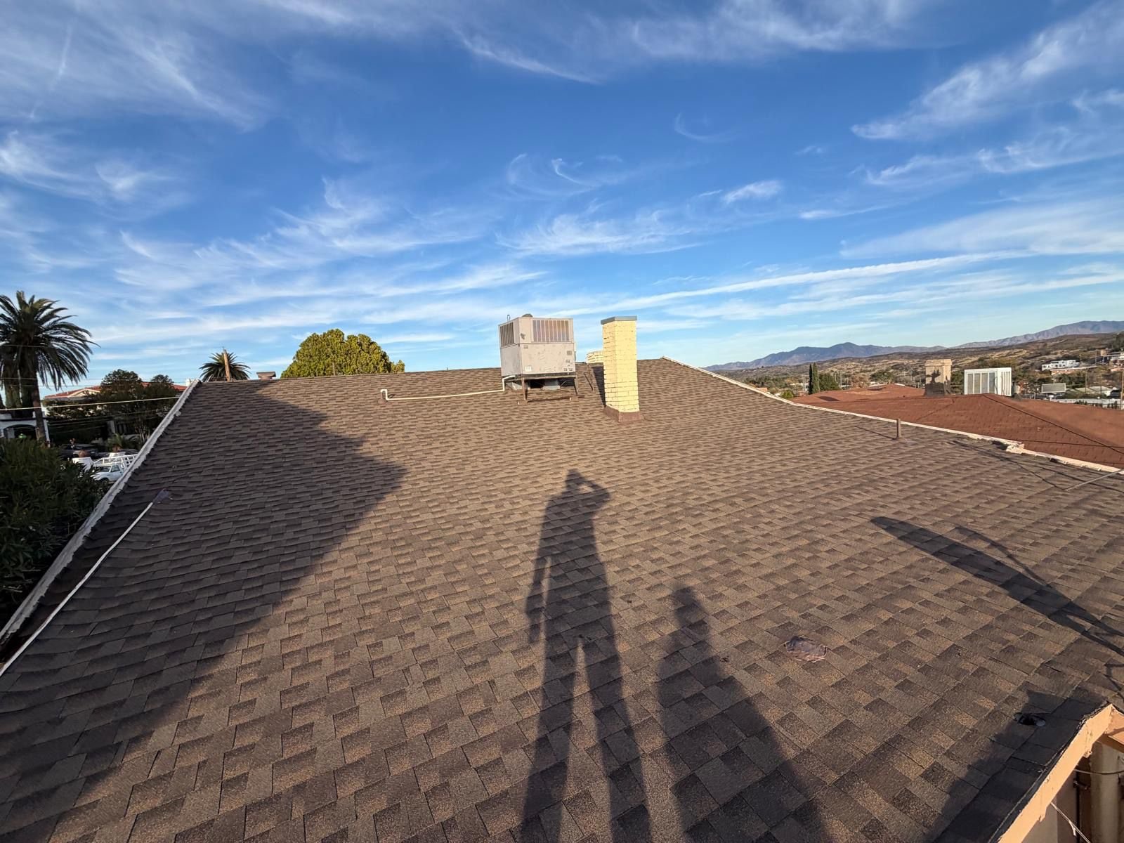 View of a rooftop with brown shingles, chimneys, and HVAC units under a blue sky with wispy clouds; shadows of two people are cast on the roof.