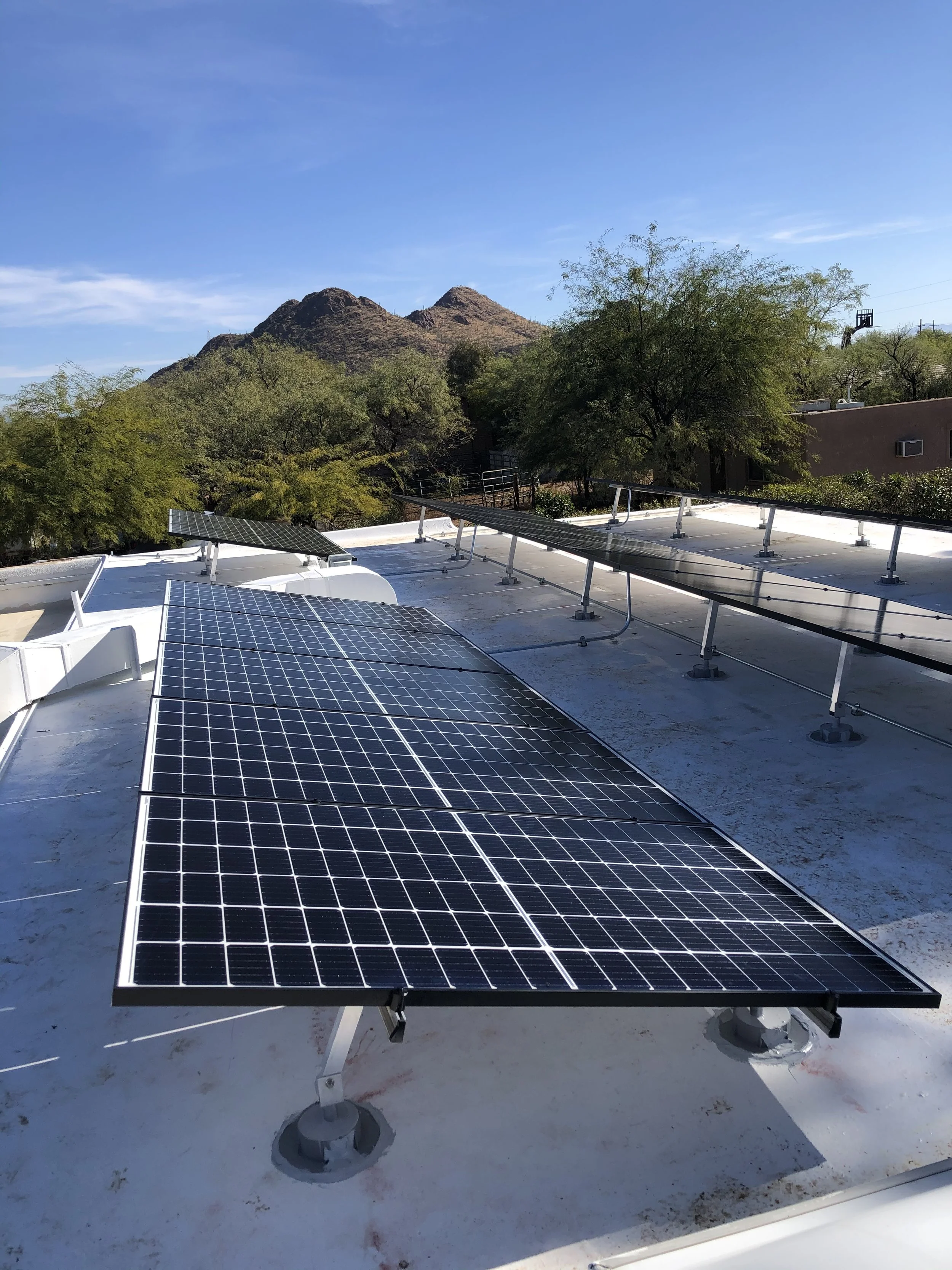 Solar panels installed on a rooftop with desert mountains and trees in the background under a clear blue sky.