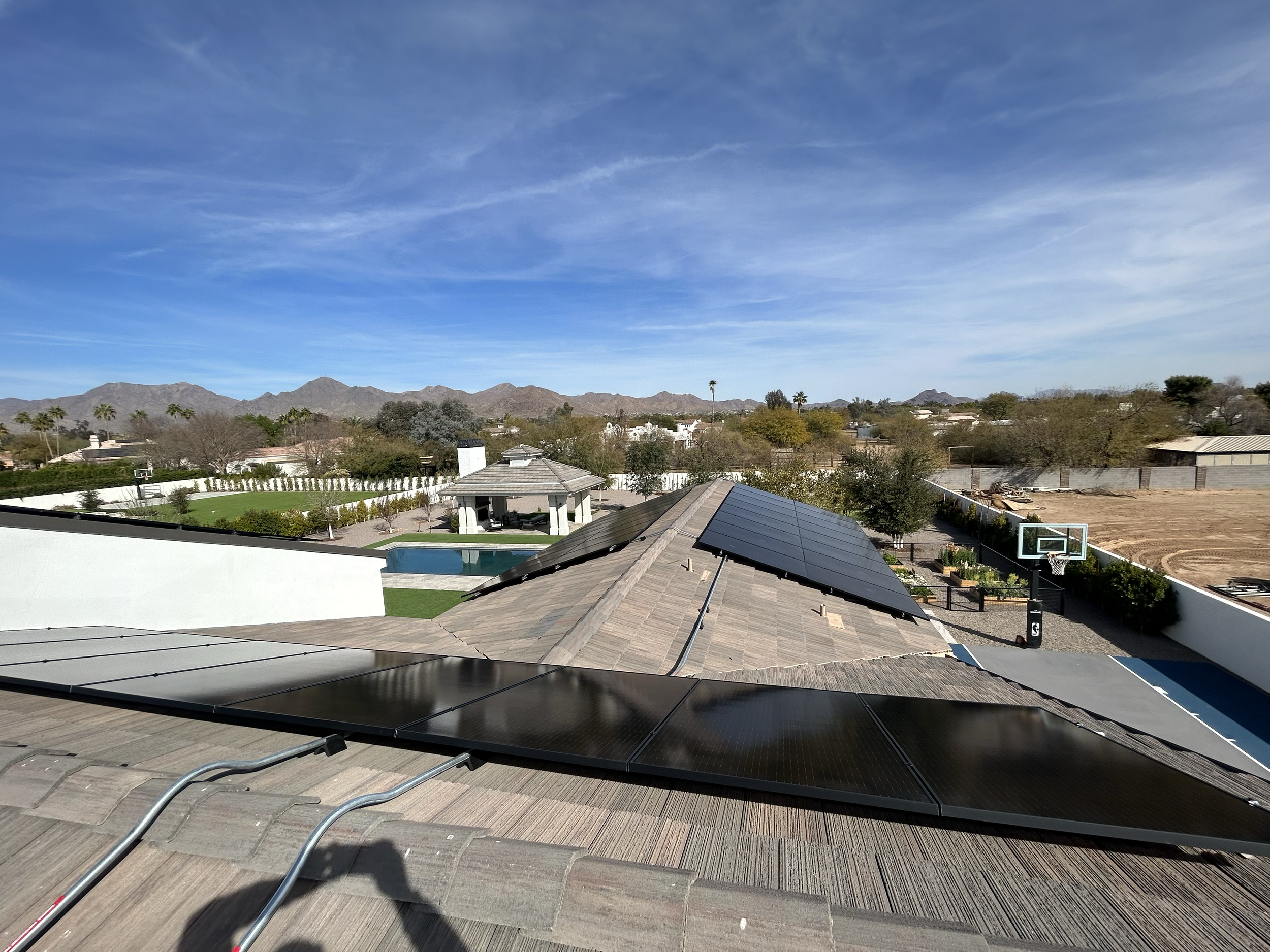 Residential backyard with solar panels installed on the roof, a basketball hoop, a swimming pool, a gazebo, and mountain range in the background under a blue sky with some clouds.