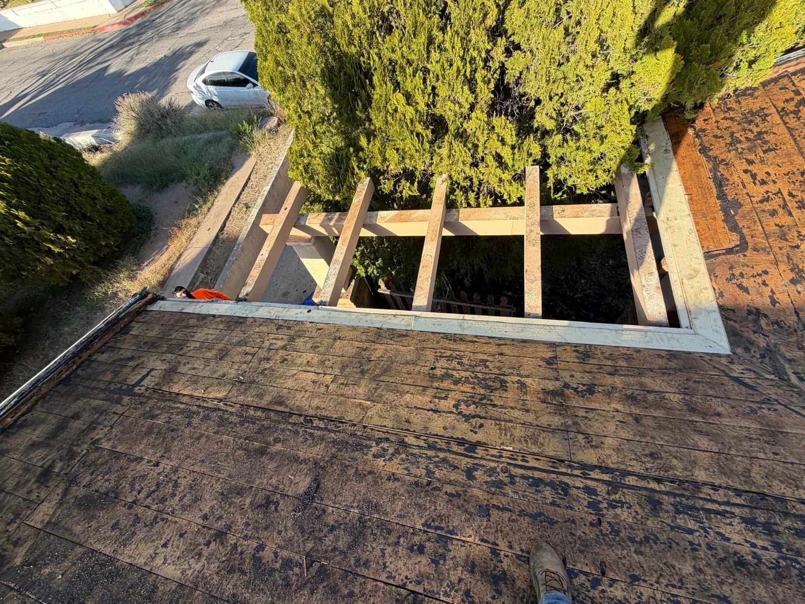 View from the top of a building showing the roof, part of the wooden structure being worked on, a person in an orange shirt, trees, and a street with a parked car.