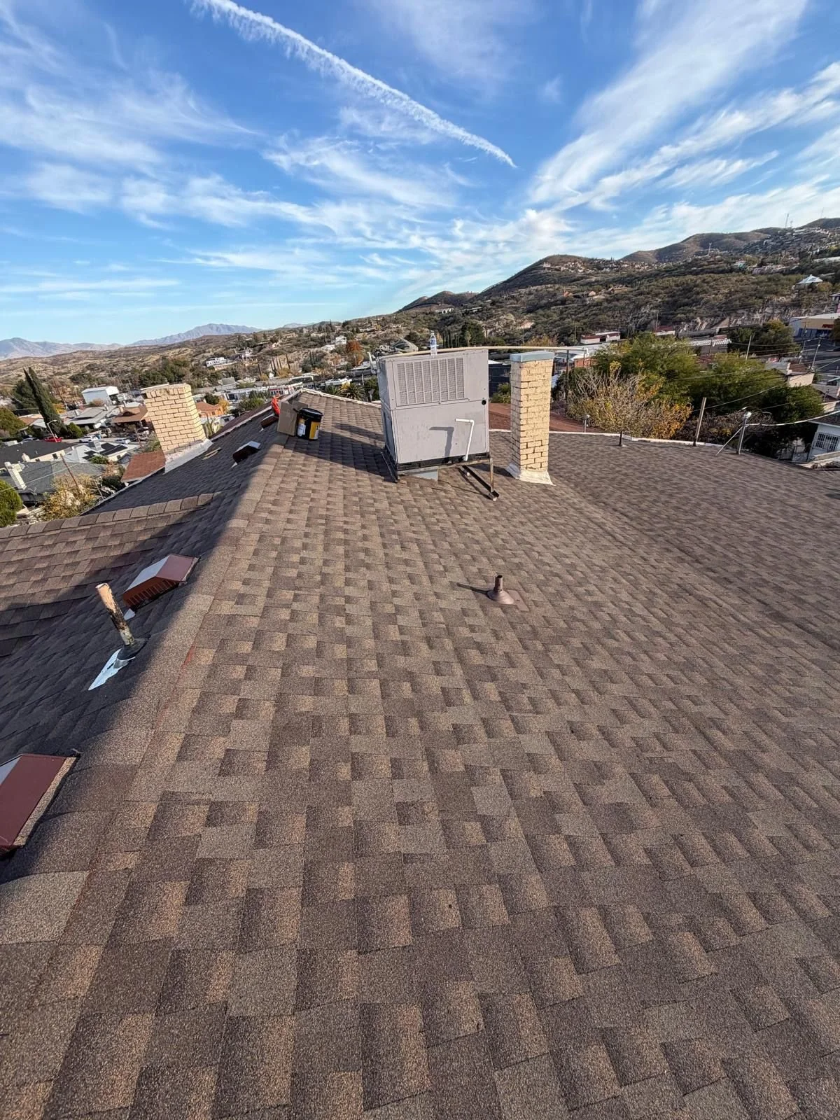 View of a rooftop with HVAC unit, chimneys, and vents, overlooking a cityscape and mountains under a blue sky with some clouds.