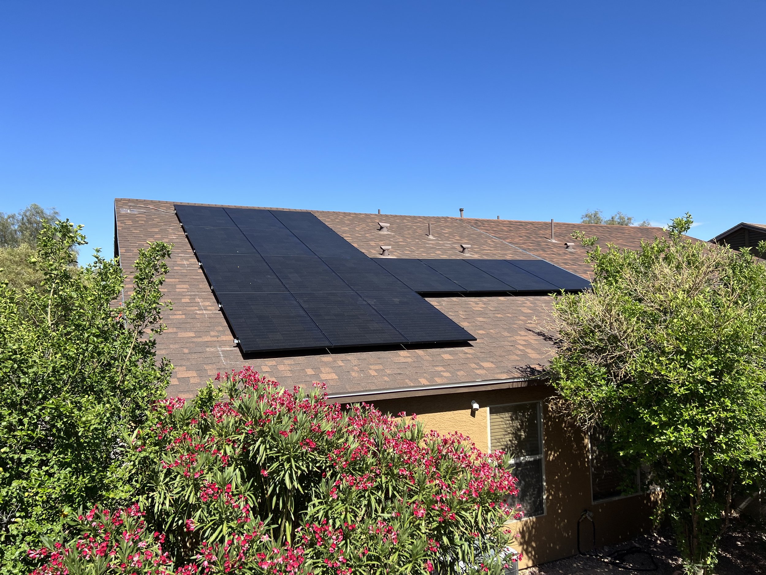 House with solar panels on the roof, surrounded by trees and flowering bushes, under a clear blue sky.