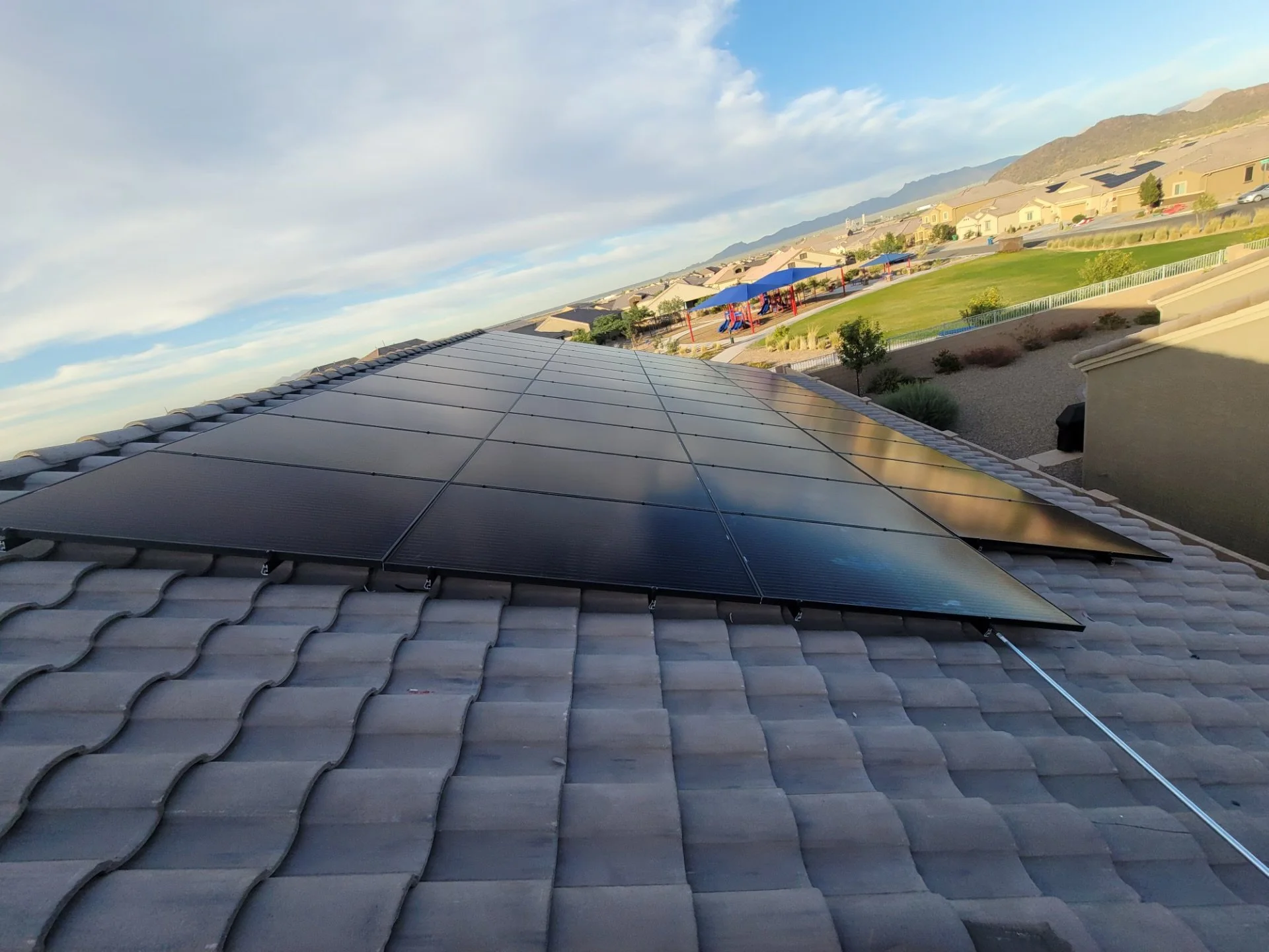 Photograph of a rooftop with black solar panels installed, overlooking a neighborhood with houses, a playground, and mountains in the distance on a partly cloudy day.