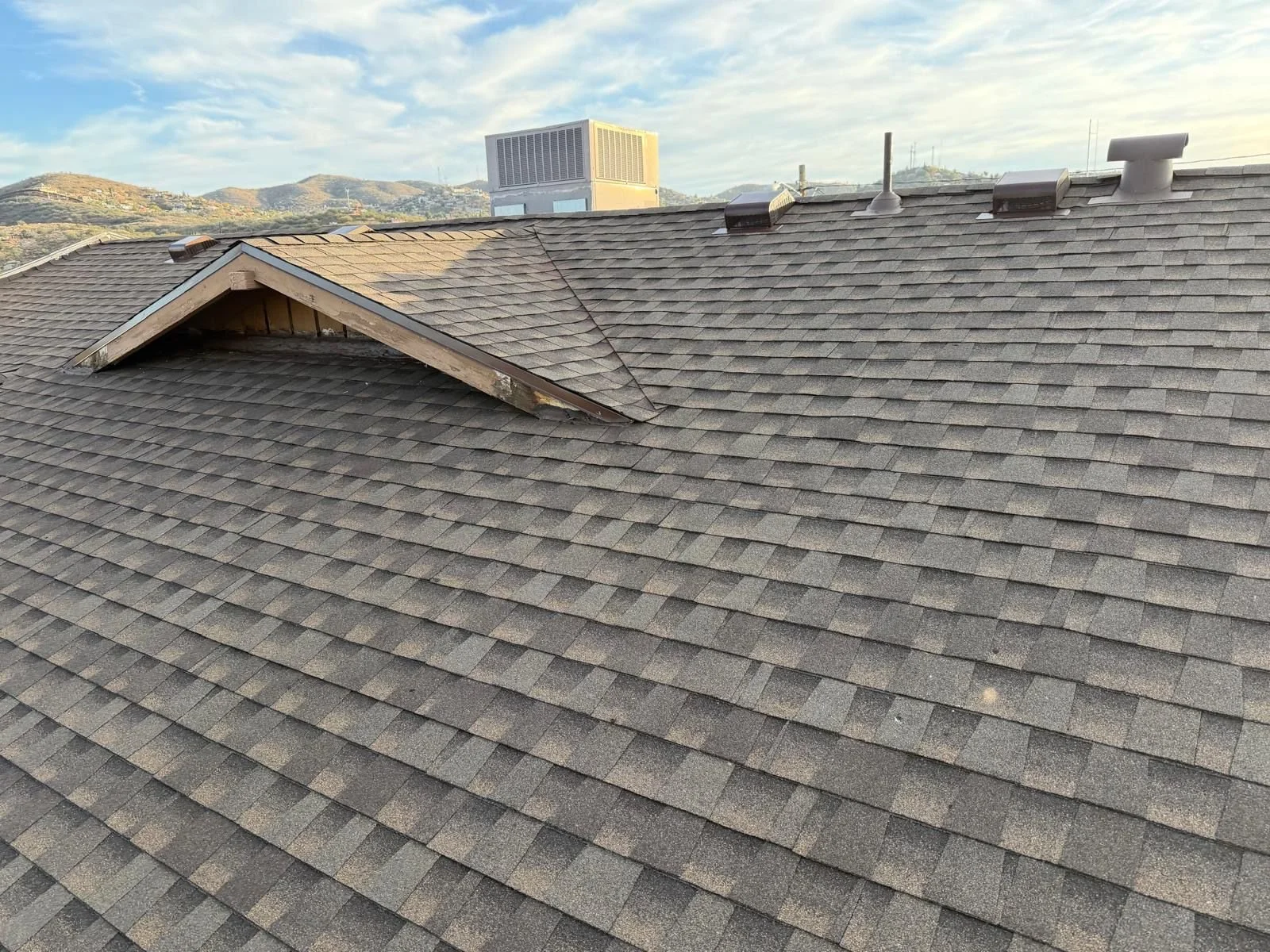 View of a building roof with new asphalt shingles, prominent gable with some damaged shingles, vent pipes, and surrounding hills in background.