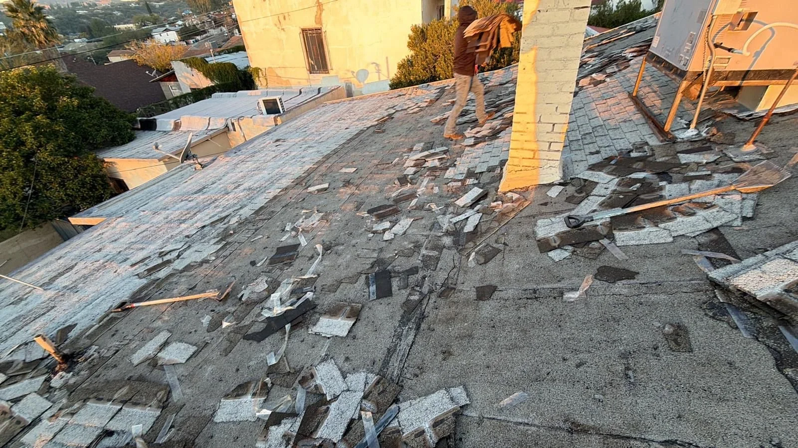 A person working on a roof with broken shingles and roofing tools, surrounded by debris.