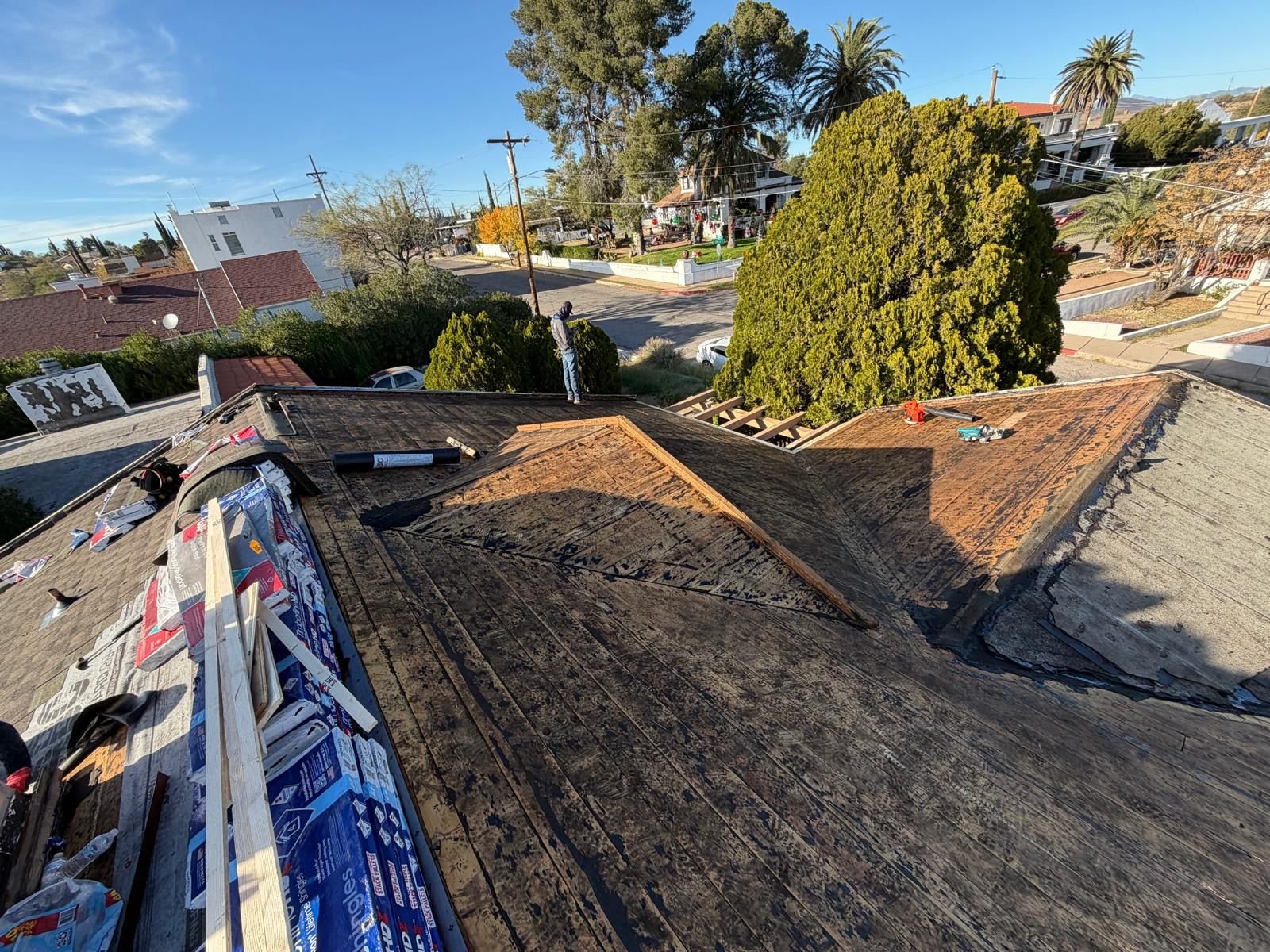 Wide-angle view of a house roof under remodeling with shingles, construction tools, and materials visible, surrounded by trees and neighborhood houses on a sunny day.