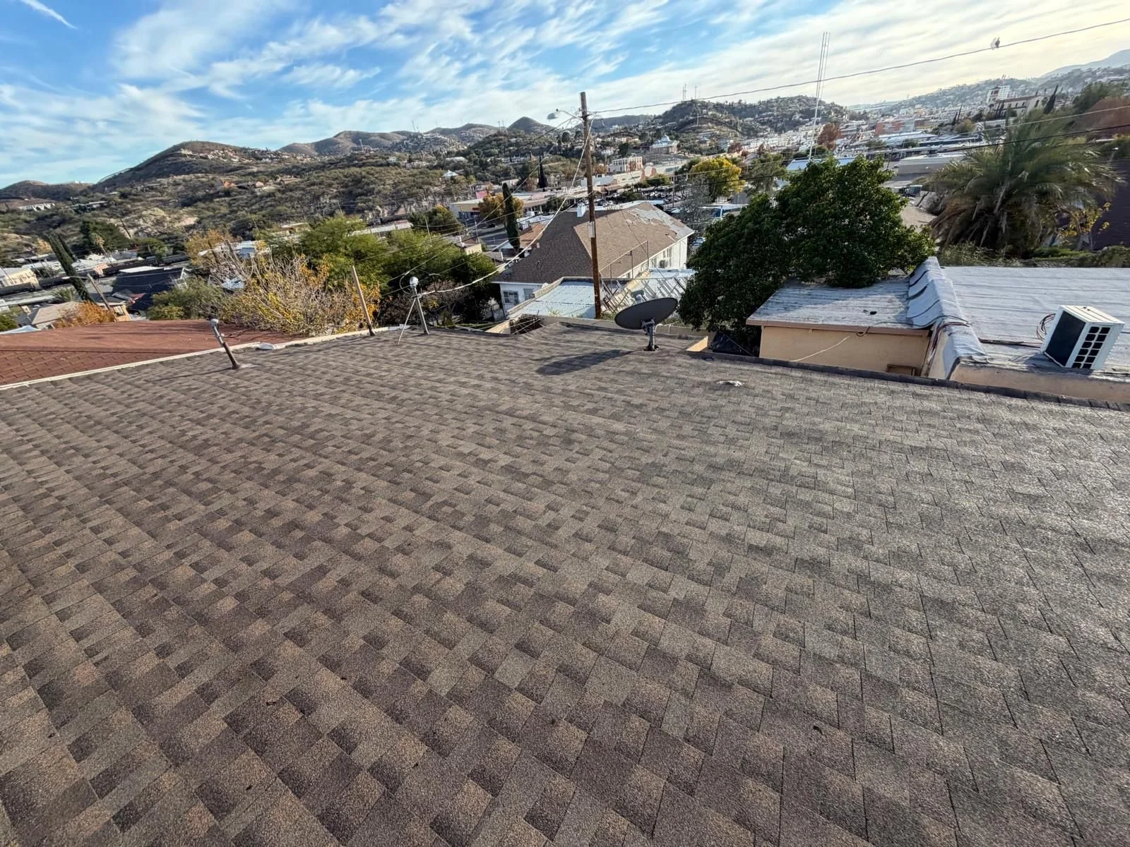 View of a rooftop with shingles, satellite dishes, and a vent, overlooking a residential neighborhood with trees, houses, and hills in the background under a partly cloudy sky.