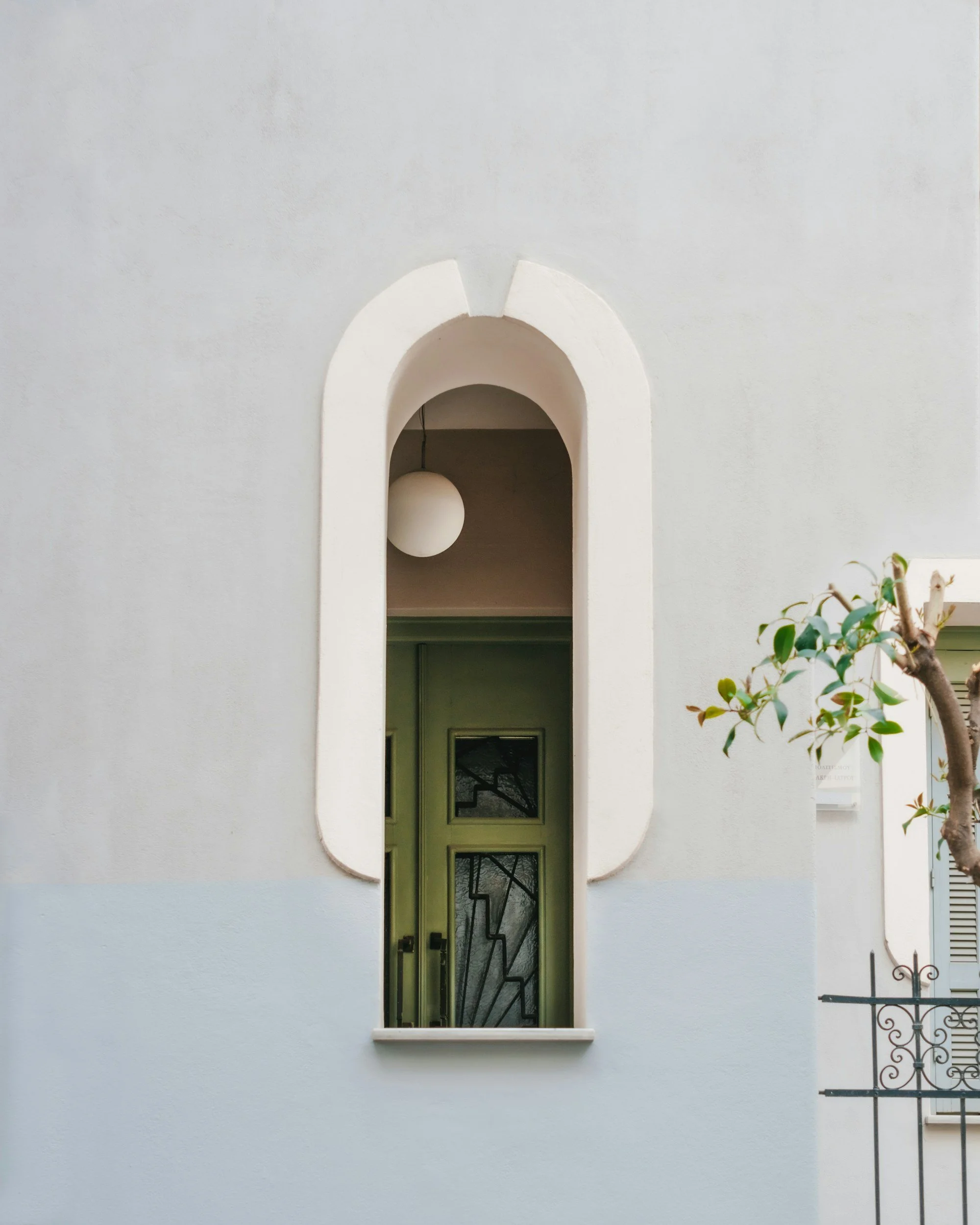 A white wall with an arched window showing a corridor with a green door and a decorative window. There's a light hanging inside the arch.