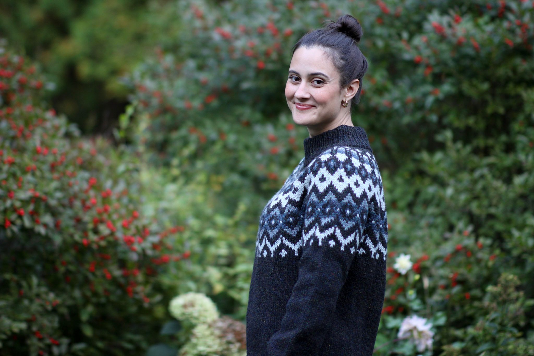 A woman with dark hair in a bun, wearing a patterned sweater, smiling and turning slightly to her left outdoors with a background of green foliage and red berries.
