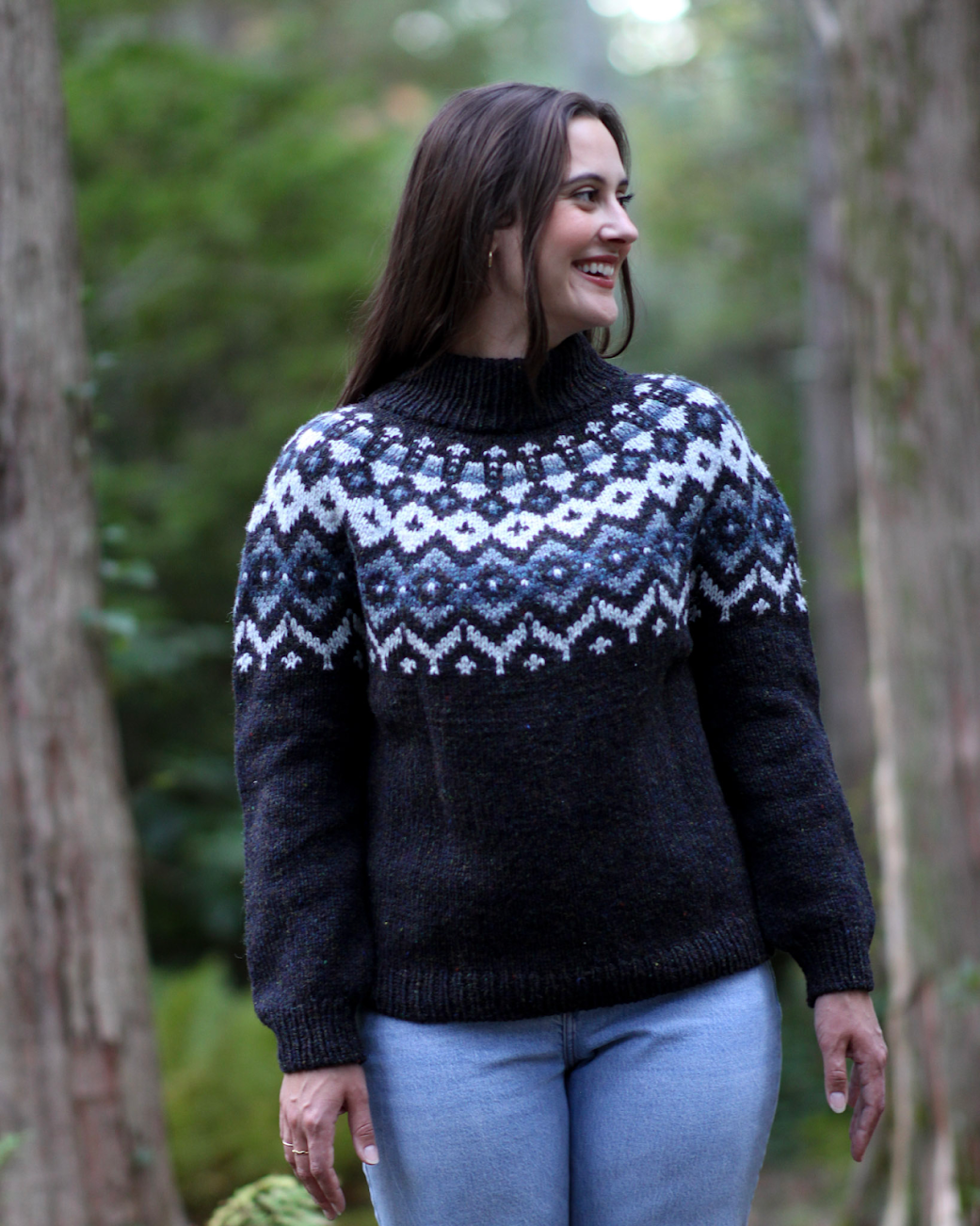 A woman with long dark hair smiling and looking to the side, wearing a black and white patterned sweater and light blue jeans, standing outdoors in a forested area.