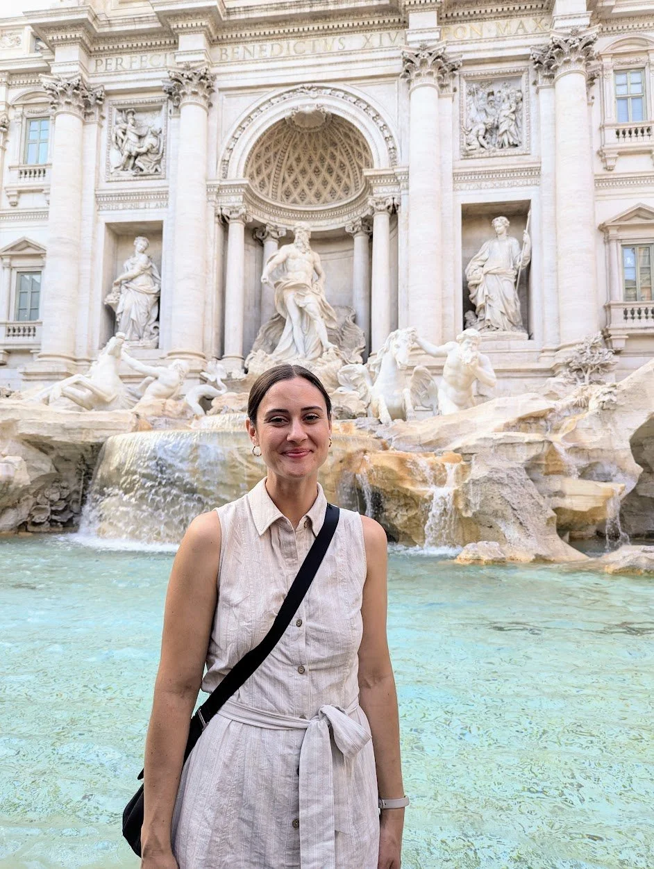 A woman standing in front of the Trevi Fountain in Rome, Italy, with a smile, wearing a sleeveless beige dress and carrying a black shoulder bag.