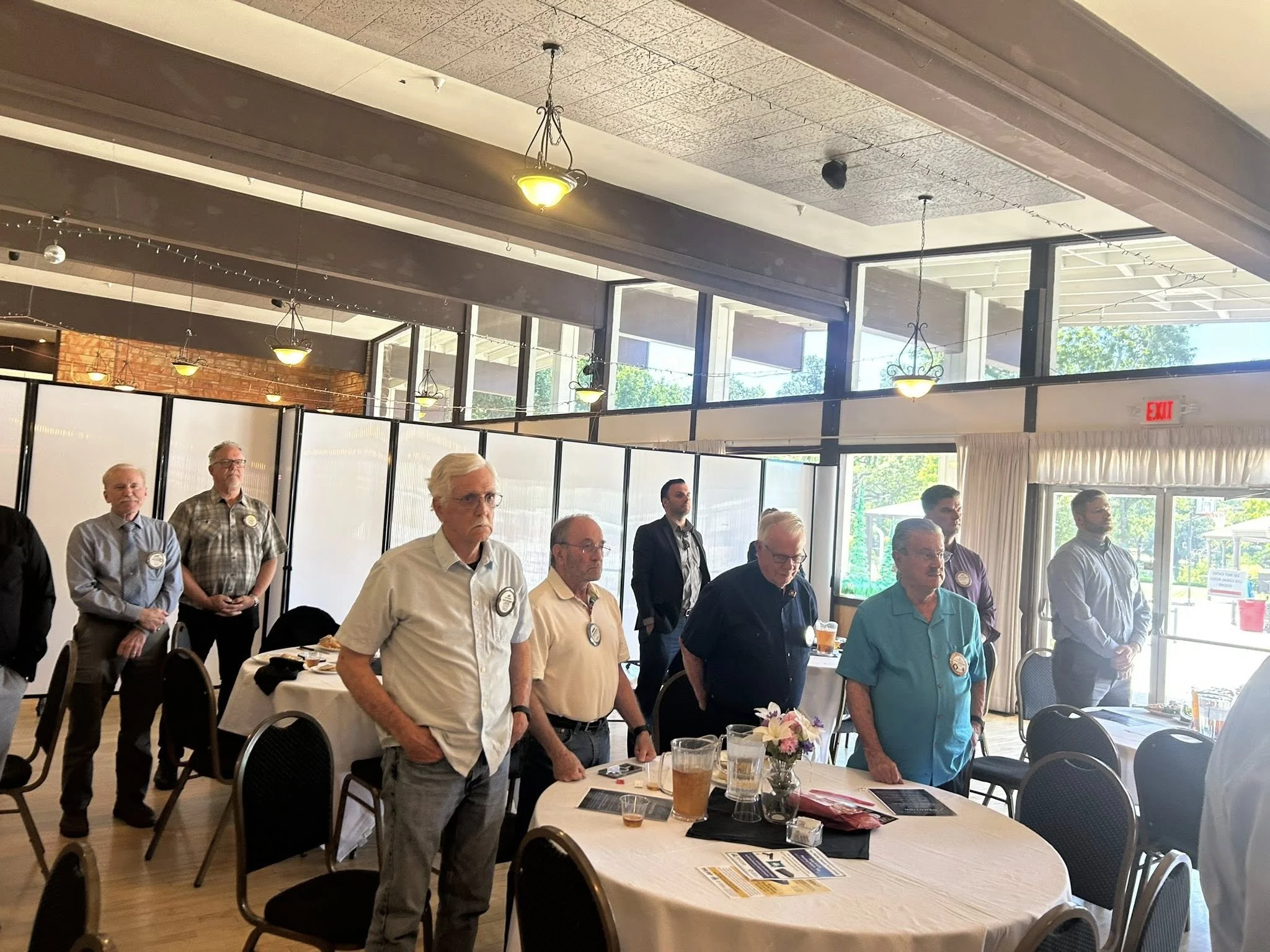 Group of men standing at a formal event in a restaurant or banquet hall, with round tables, glass windows, and hanging light fixtures.