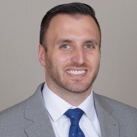 Professional portrait of a smiling man wearing a gray suit, white shirt, and blue tie against a gray background.