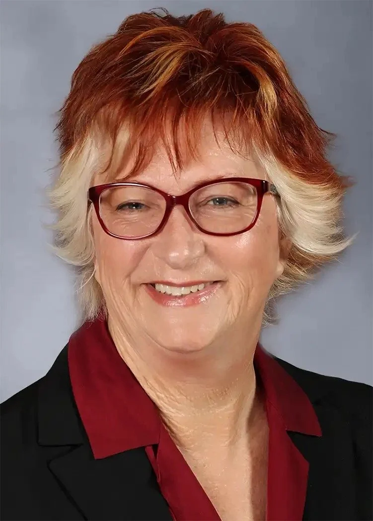 Portrait of a smiling woman with short, layered red and blonde hair, wearing glasses with red frames, a black blazer, and a red blouse.