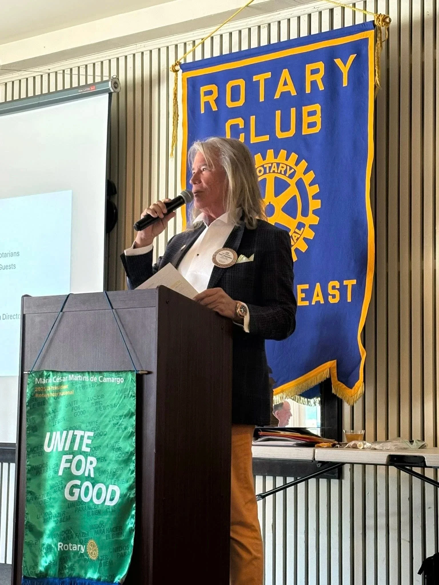 A woman with gray hair speaking into a microphone at a podium during a Rotary Club event. She is wearing a black blazer, a white shirt, and a badge. Behind her is a large blue and yellow Rotary Club banner. A green banner on the podium reads 'Unite for Good'.
