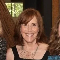 Photo of a woman with shoulder-length reddish-brown hair, smiling, wearing a black top and a necklace