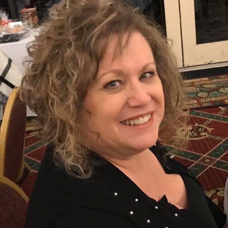 A woman with curly blonde hair smiling, wearing a black top, sitting at a table in a decorated room with patterned carpet.