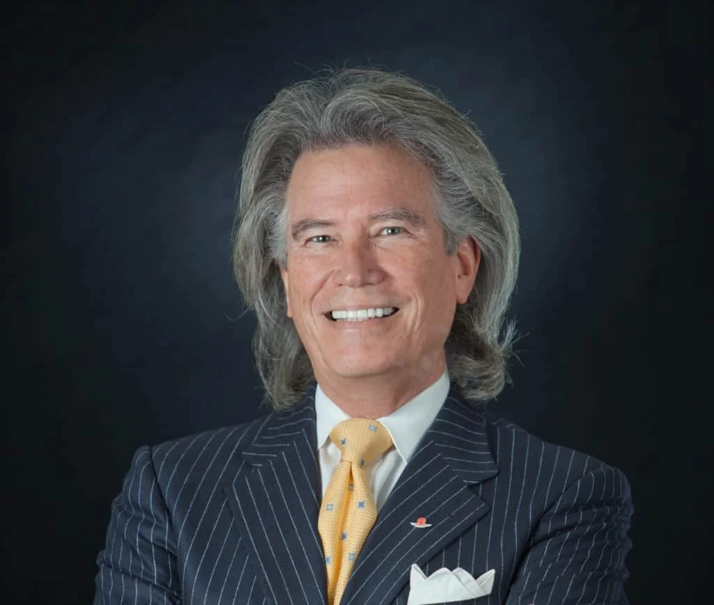 A smiling man with gray hair wearing a dark pinstripe suit, a white shirt, and a yellow tie with a small pattern, against a dark background.