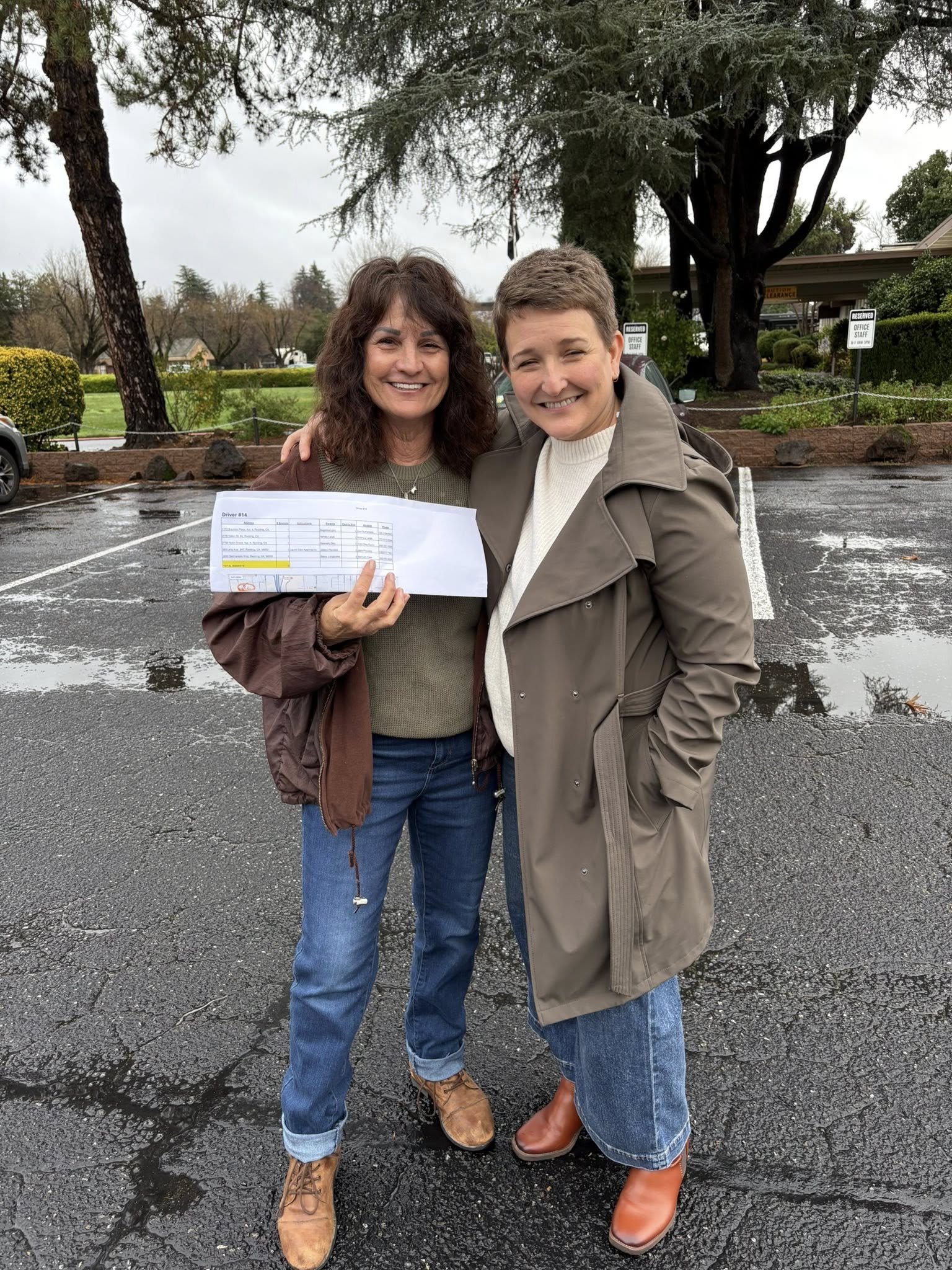 Two smiling women standing outdoors in a parking lot, one holding a document, with trees and a building in the background on a rainy day.