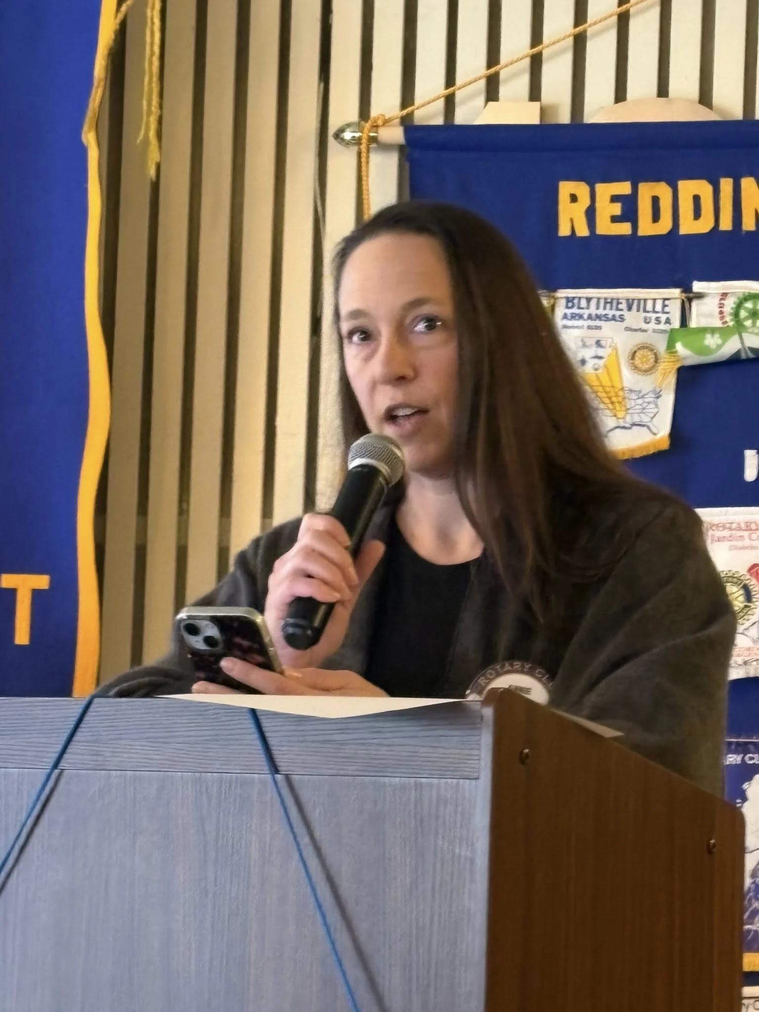 Woman with long brown hair speaking into a microphone at a podium, holding a smartphone, with a blue and yellow banner behind her.