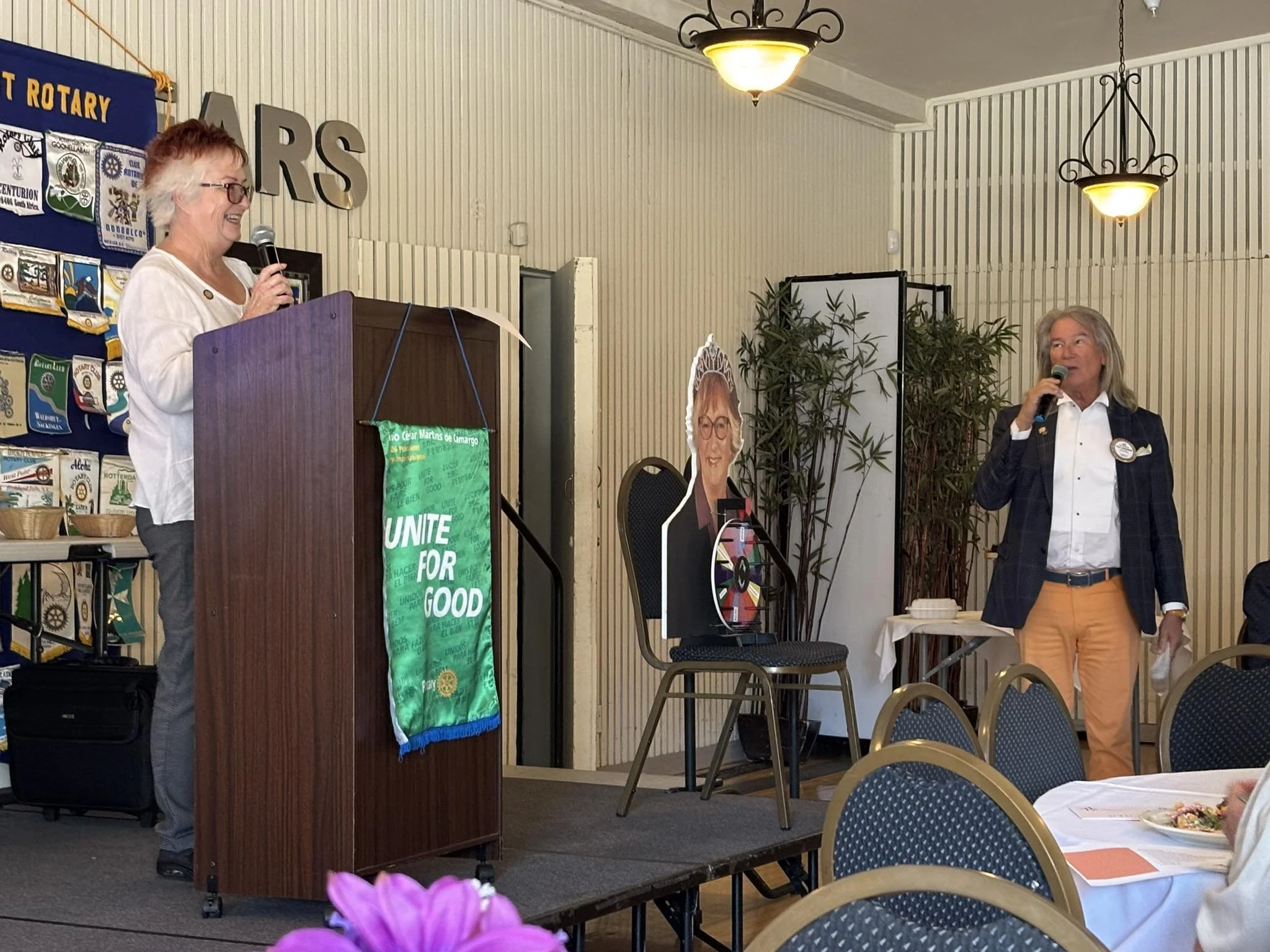 Two women speaking into microphones at a Rotary Club event. One woman stands behind a podium with a green banner that reads "UNITE FOR GOOD", the other stands to the right wearing a checkered blazer and tan pants. There are round tables with white tablecloths and chairs, a cardboard cutout of a woman, and Rotary banners on the wall.