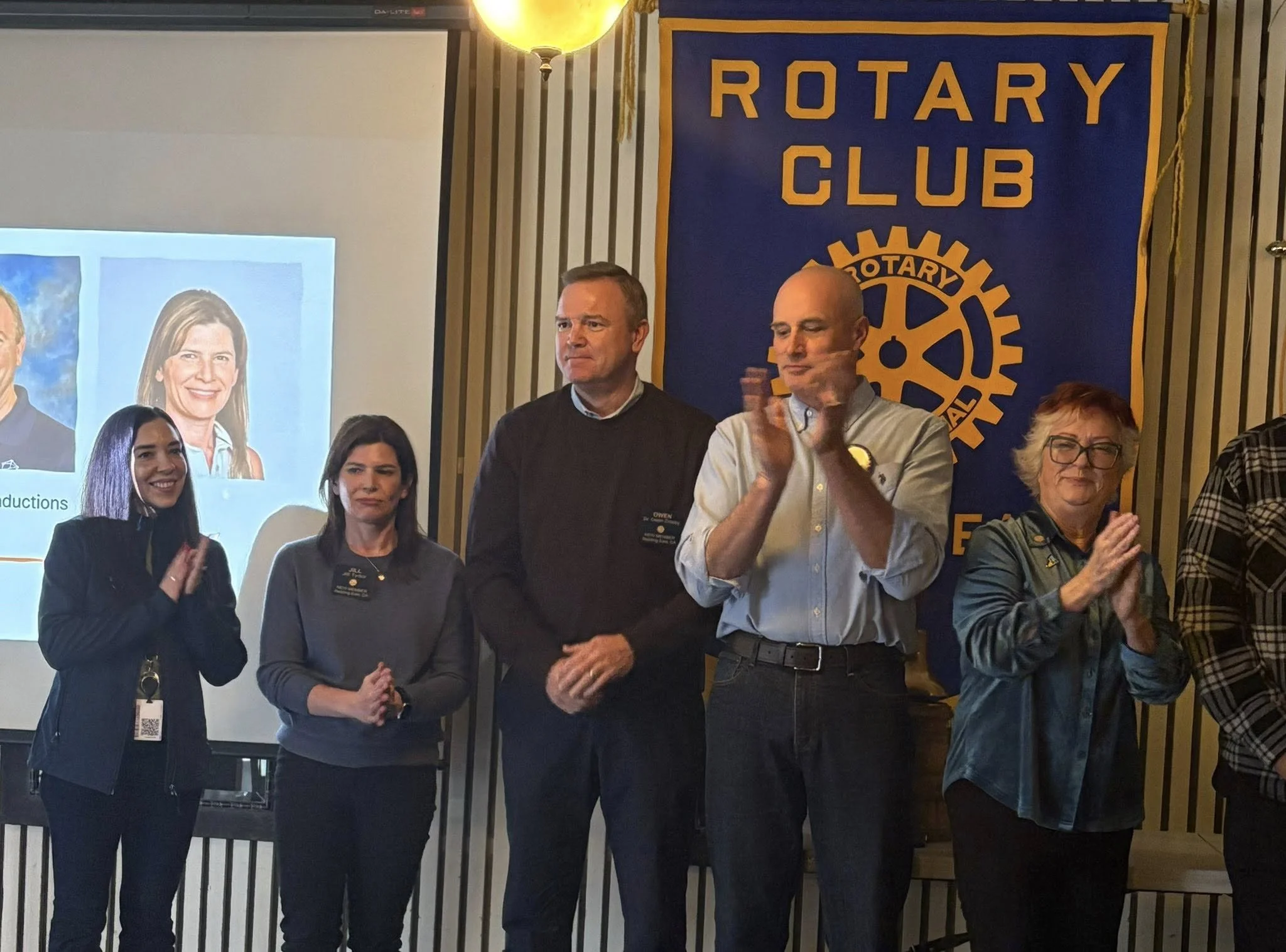 Group of five people standing in front of a Rotary Club banner, clapping, with a screen displaying a woman’s photo in the background.