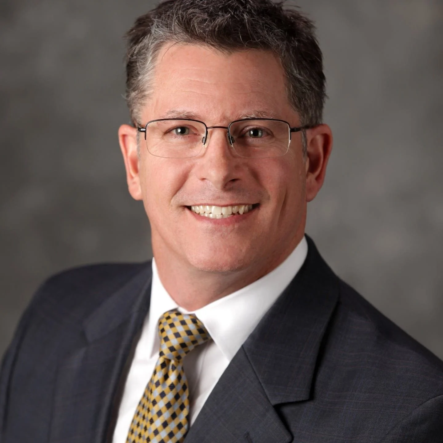 Professional headshot of a smiling man wearing glasses, a dark suit, white shirt, and a yellow patterned tie.