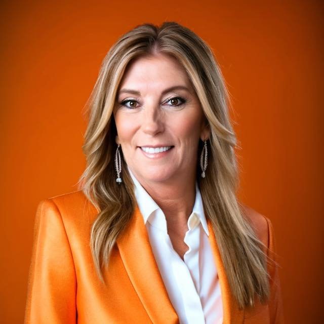 A woman with long blonde hair wearing a white shirt and orange blazer, smiling against an orange background.