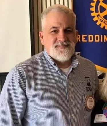 A middle-aged man with gray hair and beard wearing a striped button-up shirt standing indoors. Behind him is a rotary club banner with blue and yellow colors.